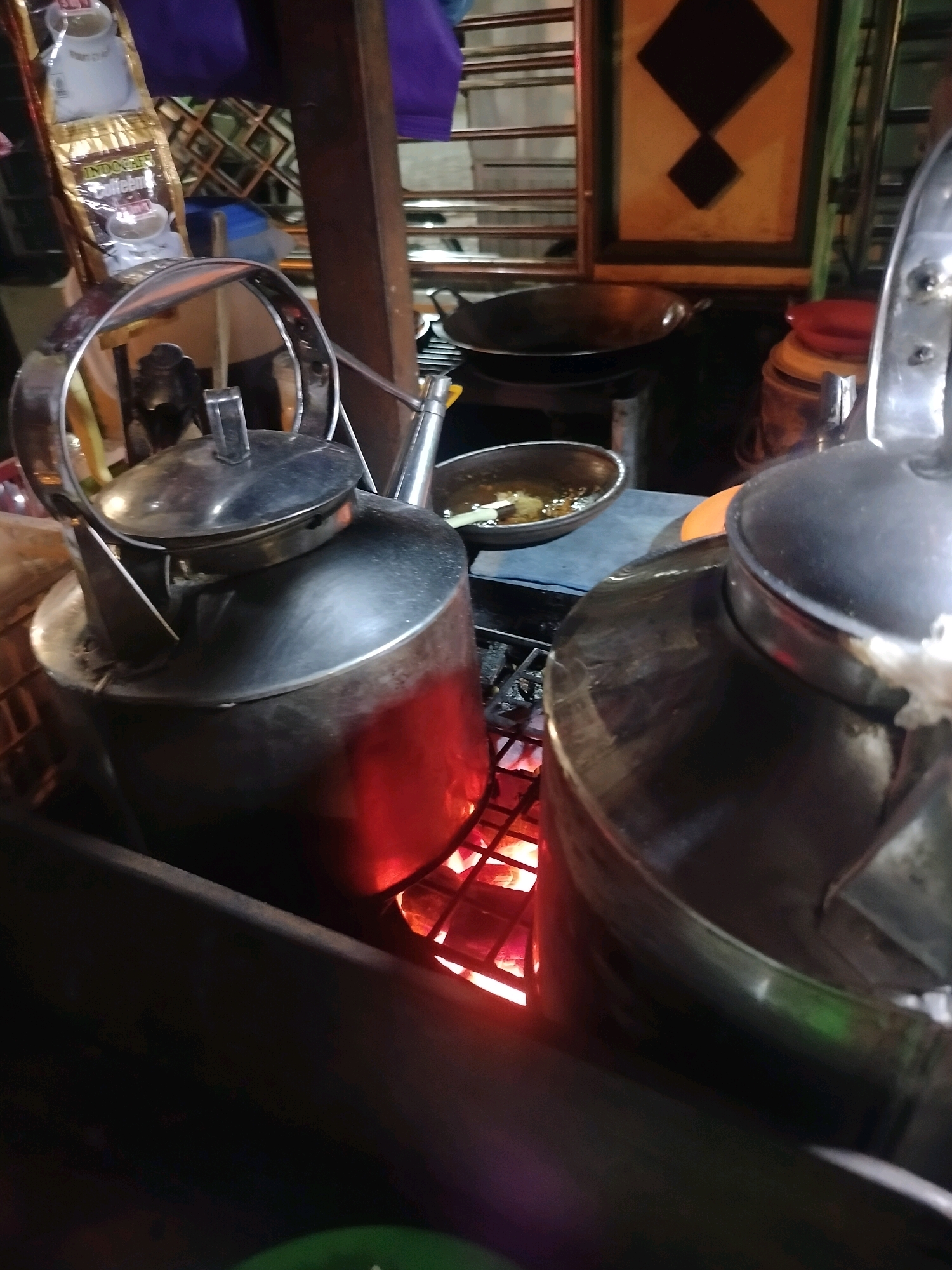This image captures an outdoor food stall in Kroya, Indonesia. The scene is dominated by two large, stainless steel kettles situated over a glowing heat source, likely charcoal, indicating that something is being heated. The kettle in the foreground is larger and positioned to the left, with a shiny metallic finish reflecting the ambient light and the red glow from below. Its lid is closed. To its right, another, partially visible kettle is also present.
In the background, there are various elements that suggest a functional food stall. A metal wok is placed on a stand to the left of the kettles. A small bowl with some liquid and what appears to be food is visible on a blue surface. To the far left, a golden packet of "Indocafe" coffee is hanging, suggesting that coffee is also being sold or prepared. The stall structure appears to be made of wood. The lighting is dim, with the prominent red glow from the heat source casting warm tones on the kettles and surrounding objects. The overall environment suggests a bustling, potentially nocturnal, street food setting.