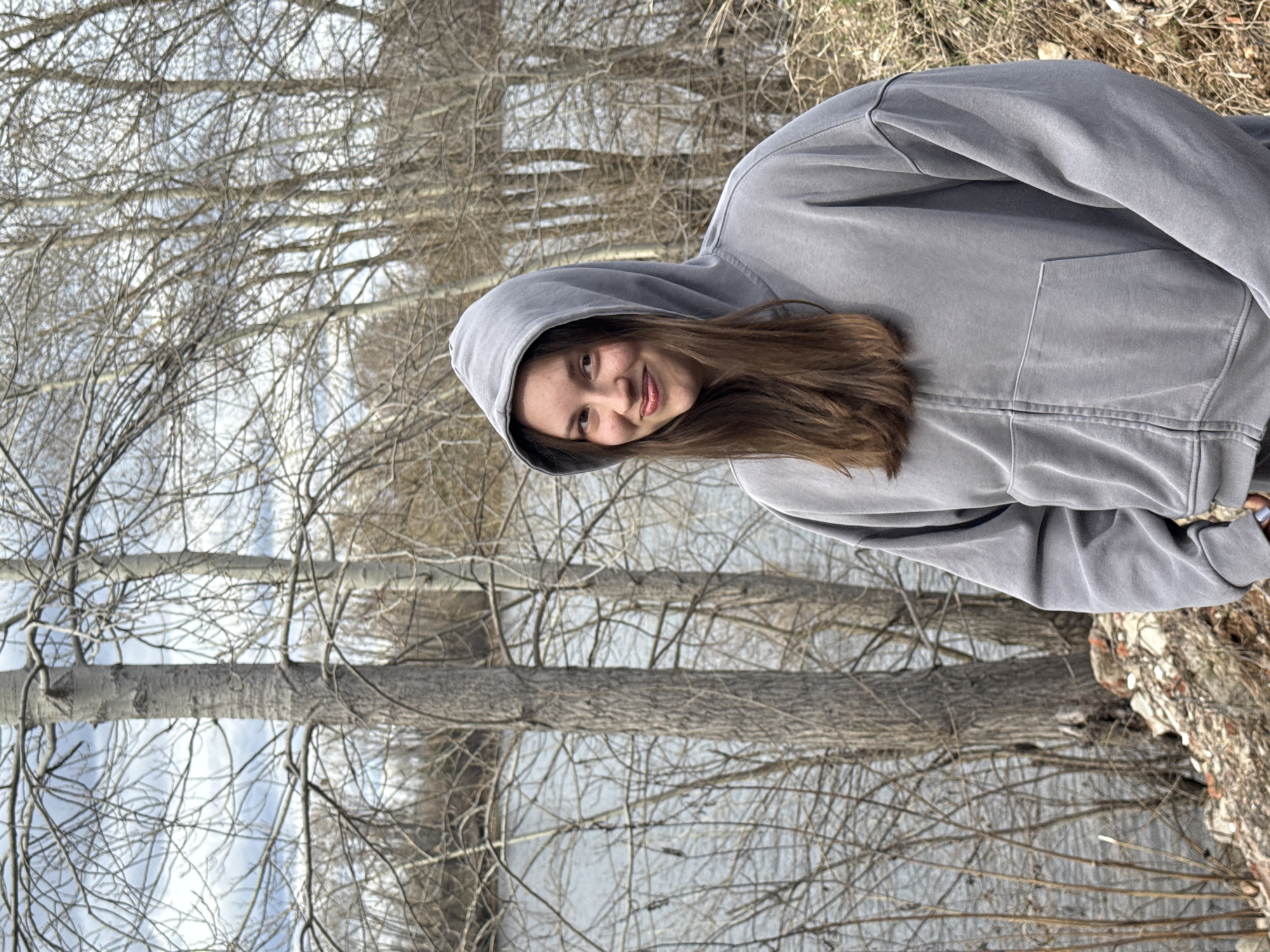 A photograph features a person wearing a grey hooded sweatshirt posing against a backdrop of bare trees and what appears to be a body of water, possibly a river. The location may be Chaikovsky, Russia, though this cannot be definitively confirmed from the image alone. The trees are leafless, indicating a winter or early spring season. The person is looking at the camera, with a slight smile.