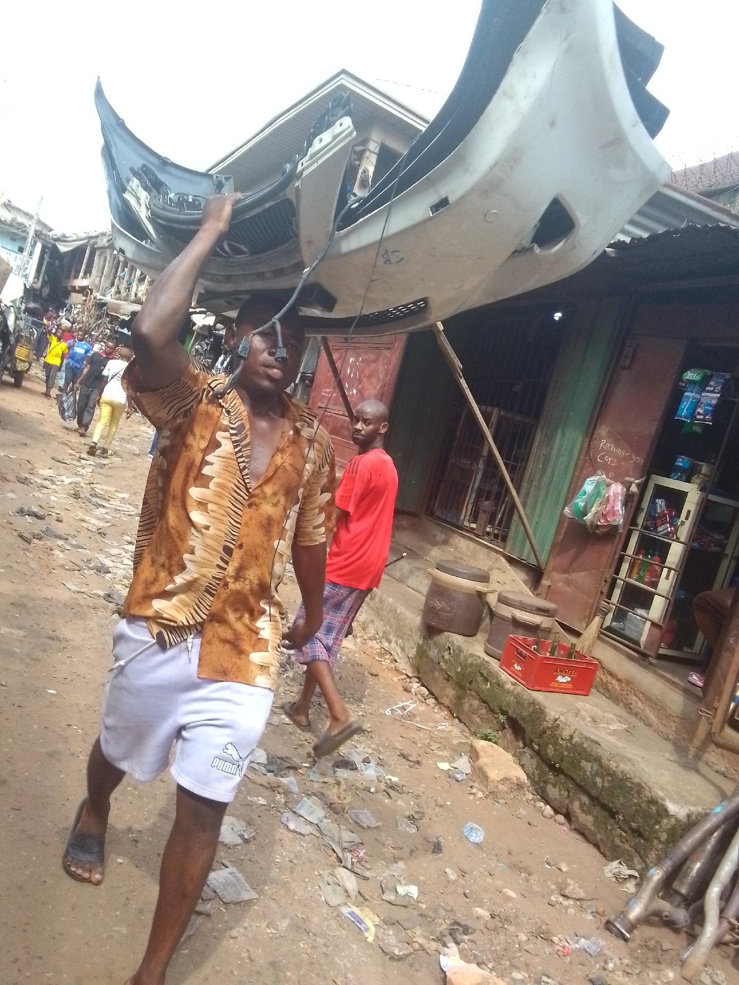 This image depicts a bustling street scene in Onitsha, Nigeria, under natural daylight.
The central subject is a man wearing a brown patterned short-sleeved shirt and white shorts with a "PUMA" logo. He is carrying a large, light-colored car bumper, which appears to be a front bumper with black grille elements and attached wiring, balanced on his head and right shoulder. He looks directly at the camera with a neutral expression. He is wearing dark sandals.
Slightly behind and to his right stands another man wearing a red t-shirt and plaid shorts, also in sandals. He is looking back over his shoulder.
The setting is an unpaved, dusty street littered with debris. Along the right side, there are simple shopfronts constructed with corrugated metal, some with closed metal doors and others partially open. One shop on the right has an open display case stocked with packaged goods. Outside this shop, two brown earthenware containers sit on a concrete ledge next to a red plastic crate branded "AMSTEL MALTA" containing bottles. Faint writing on the shop wall reads "Recharge your" and "Card." In the background, numerous other people are visible walking along the crowded street, suggesting a busy market or residential area. The overall atmosphere is one of active commerce and daily life.