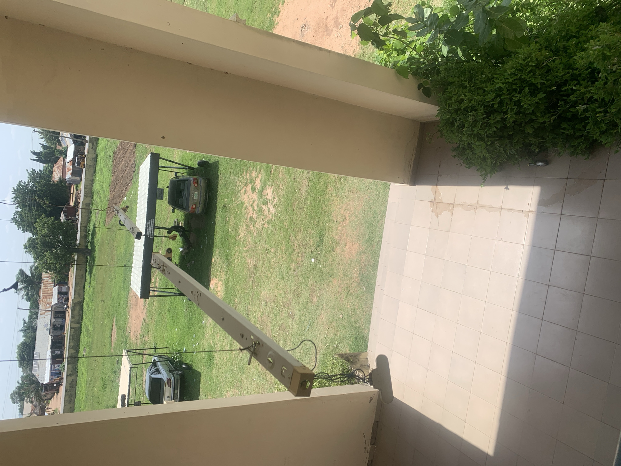 The image captures a scene in Jalingo, Nigeria, likely within an educational or governmental institution, given the presence of a covered parking area and a sign that reads "DEAN". Two cars are parked under a white-roofed structure. A silver sedan is parked in the foreground under one of these shelters, and another silver sedan is visible further back. A person is seen working under the shelter near the second car. In the middle ground, a long, rectangular fluorescent light fixture hangs suspended from wires, positioned horizontally above the ground. This fixture appears to be either undergoing maintenance or is in the process of being installed or removed, as cables are connected to it. Beyond the covered area, a grassy field stretches out, with some areas showing tilled earth, possibly for agriculture or landscaping. In the background, a row of buildings with corrugated iron roofs line a street, suggesting a commercial or residential area. Trees dot the landscape, and the sky is partially visible, indicating daytime. The foreground shows a tiled patio area, with a large beige pillar on the right and another on the left, framing the view. Lush green bushes are growing near the pillar on the right.