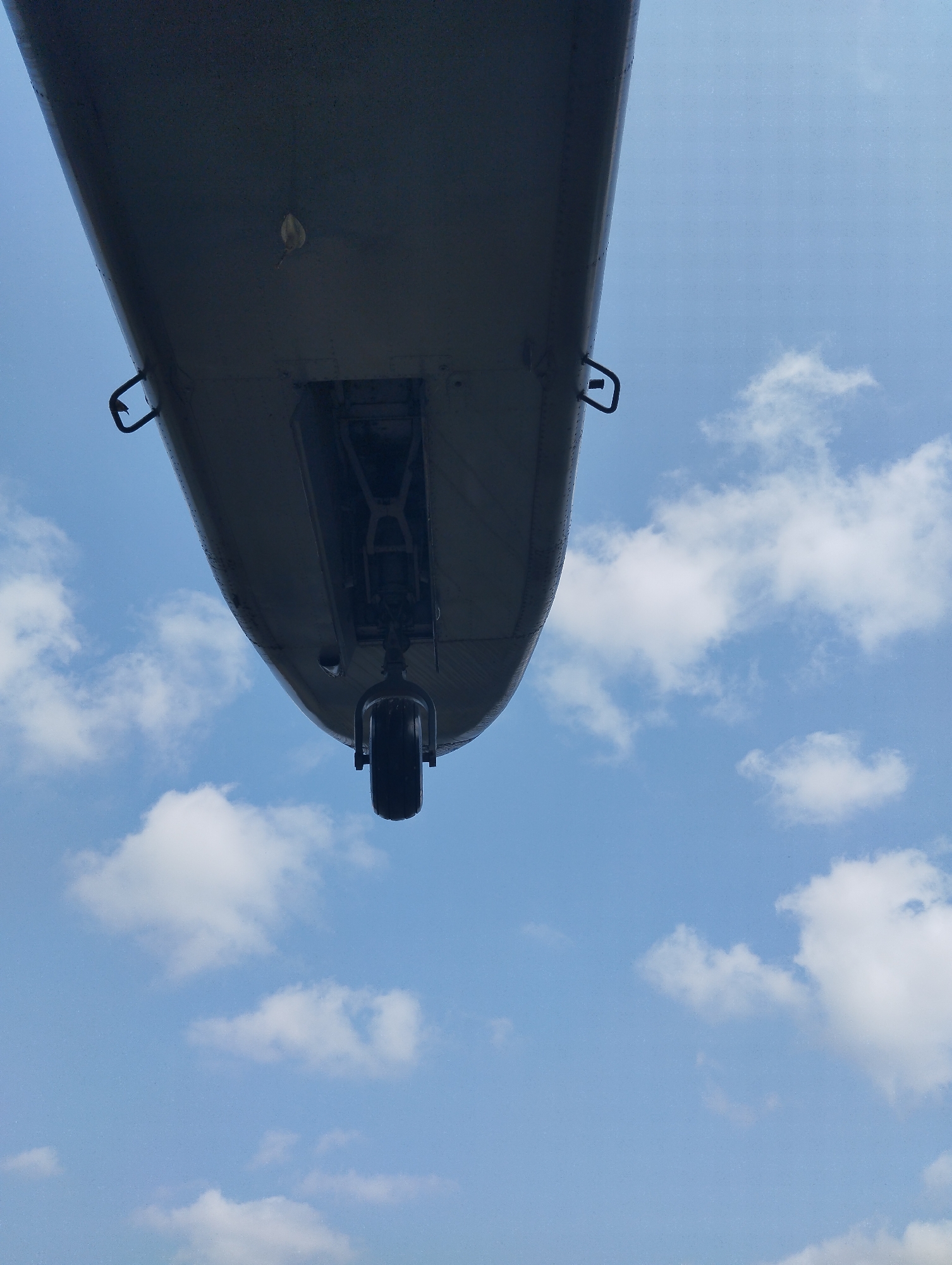 This is a low-angle, close-up, full-frame photograph of the underside of what appears to be an aircraft's wing and a visible landing gear assembly with a single wheel. The wing is a dark, metallic gray, showing a smooth, curved surface with a line of rivets running along its edge. There are two small, dark, loop-like structures attached to the underside of the wing, possibly for securing covers or equipment. A darker, more complex mechanical assembly is visible within a recessed section of the wing, leading to the main landing gear strut and wheel. The wheel is black and circular, with a portion of its tire visible.
The background is a bright, clear sky with scattered white, fluffy cumulus clouds. The lighting suggests it is daytime, with the sun likely positioned above and to the side of the frame, casting subtle shadows on the underside of the wing. There is no indication of the location being in Betro, Indonesia. The image solely depicts an aircraft component against a sky.