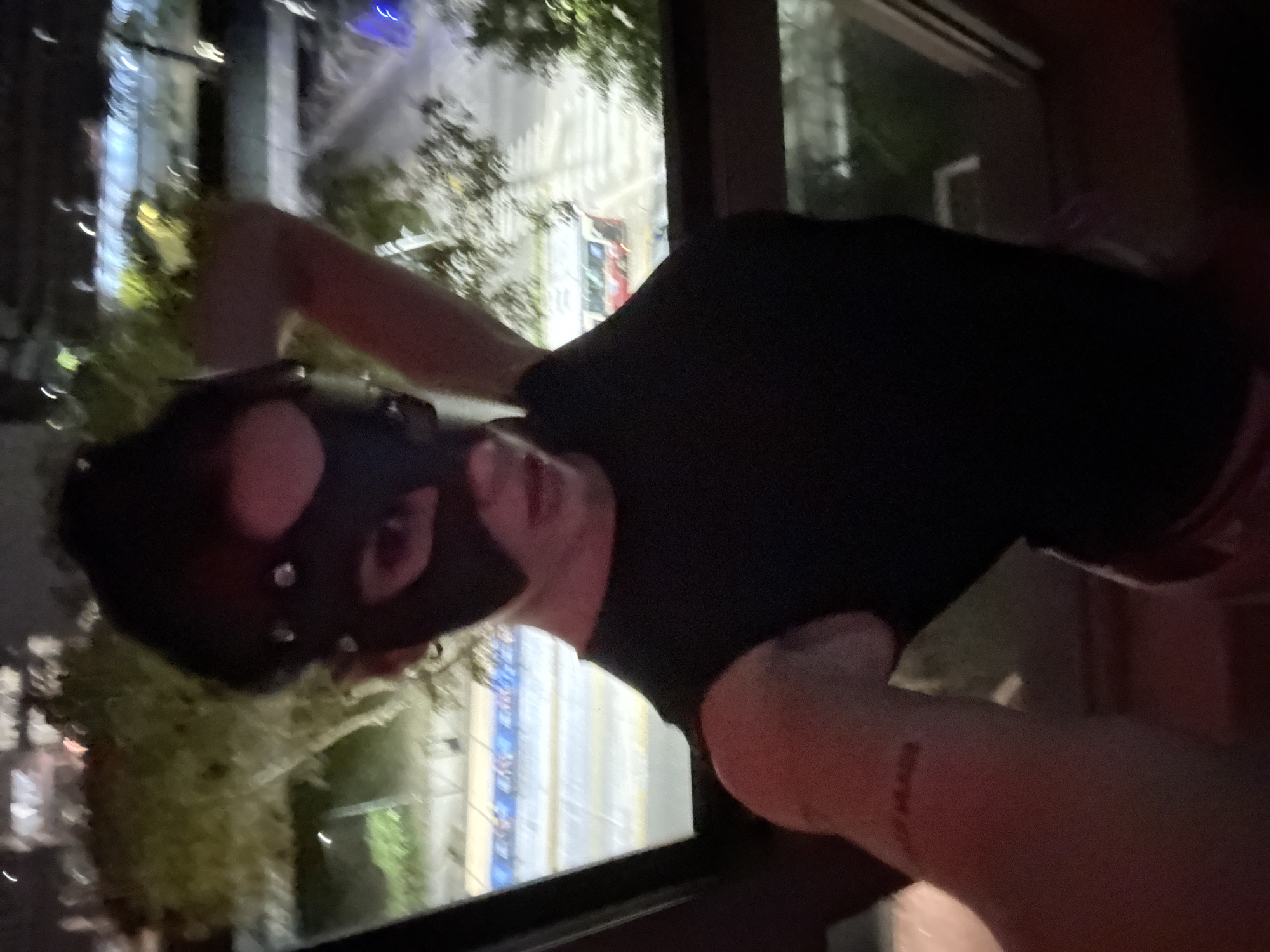 The image features a person indoors at night, positioned to look out a window at a blurred urban landscape. The individual wears a black mask, possibly a pup play hood, which covers the entire head and face, featuring cutouts for the eyes and mouth, and is adorned with small metallic studs. They are also wearing a black t-shirt or top.
Their left arm is visible, bent at the elbow, with a notable reddish marking on the inner forearm. A closer examination of the crops reveals this marking to be text, clearly spelling "BUENOS" in a stylized font, likely a tattoo or temporary body art. This detail directly ties to the provided location context of Buenos Aires, Argentina.
Outside the window, the scene is dark, indicative of nighttime, with various artificial lights creating blurs and reflections on the glass. Hints of tall buildings, vertical light sources, and green foliage are discernible through the reflections and blur. One crop shows a plant very close to the window, possibly on a balcony. The person is actively posing for the camera, looking towards the lens. The environment appears to be an urban setting, observed from an indoor vantage point.