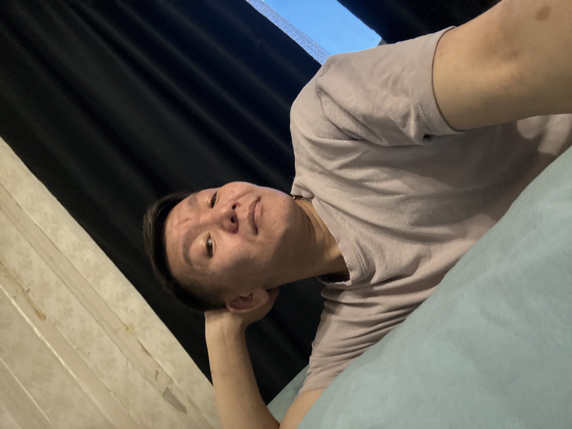 In Khimki, Russia, a person is lying down, resting their head on their arm. They are wearing a light purple t-shirt and appear to be indoors, possibly in a bedroom. The background features black curtains and a portion of a light-colored textured wall. The lighting suggests it might be evening or a dimly lit room. The person's gaze is directed slightly upwards.