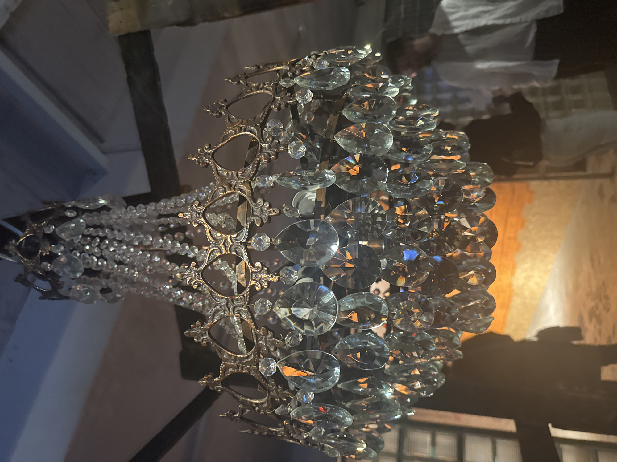 The image shows a close-up, angled view of an ornate crystal chandelier. The chandelier is elaborate, featuring multiple tiers of faceted crystals that refract light, creating a dazzling effect. Some of the crystals are large and teardrop-shaped, while others are smaller and round. The metal framework of the chandelier is detailed with intricate scrollwork and decorative elements, appearing to be a dark, antique bronze or brass.
The background is dark and somewhat out of focus, suggesting an interior setting, possibly a room with ambient lighting. There are hints of a ceiling or structural beams at the top of the image. In the mid-ground, a blurred area with warm, orange and yellow tones suggests a light source or decorative element, possibly a lamp or wall sconce, casting a glow. Further back, a shadowy, indistinct shape could be furniture or another object in the room.
The lighting in the image is dramatic, with the chandelier's crystals reflecting and amplifying the light. The overall mood is one of elegance and luxury. There is no visible text in the image. The context of Moscow, Russia, doesn't provide specific visual cues in this close-up of an object, but implies a potential setting within a historical building, hotel, or upscale residence.