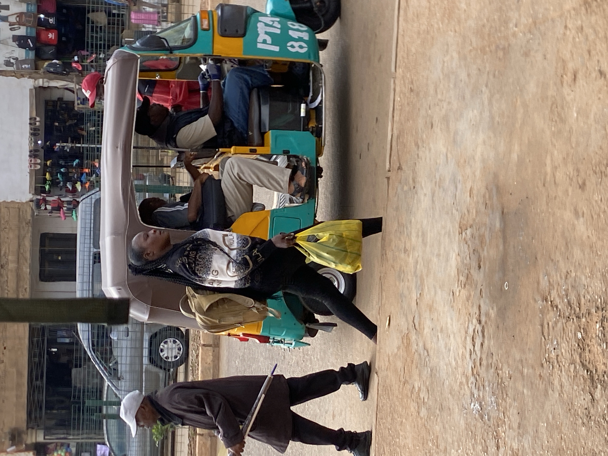 In Bukuru, Nigeria, a woman with braided hair carries a yellow plastic bag and a backpack while walking past a three-wheeled auto-rickshaw. The auto-rickshaw, painted teal and yellow, has "PTA 818" written on its side. Inside, a man wearing a red cap drives, and another man sits next to him. A man in a brown coat and a white cap walks in the foreground, holding a long object. The street is unpaved, and buildings line the background.