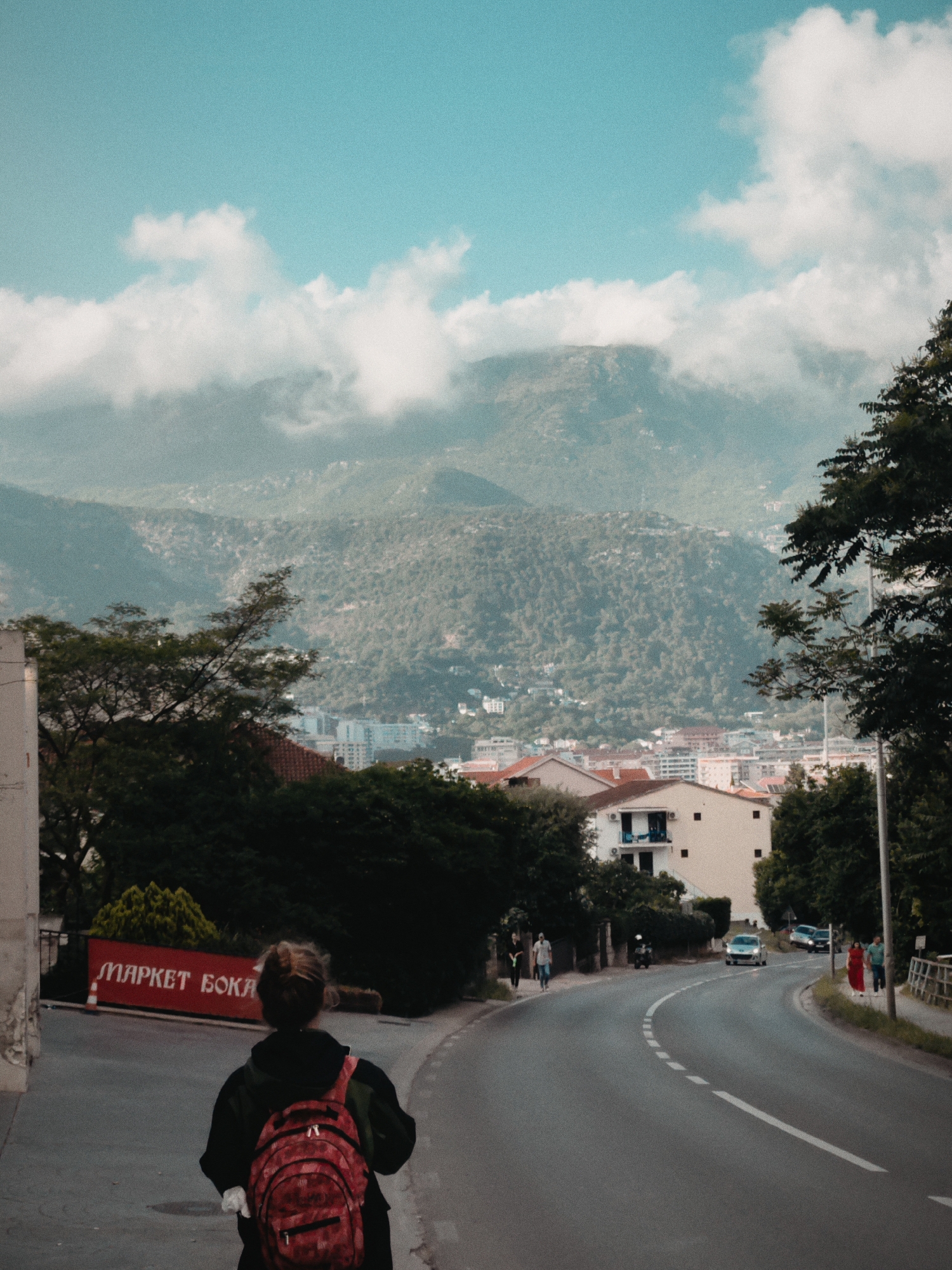The image depicts a view of Budva, Montenegro, looking towards mountains partially obscured by clouds. In the foreground, a person with a red backpack walks down a paved road. A sign reading "MAPKET БОКА" is visible to the left. The road curves past houses and trees toward the city below and mountains in the distance. Cars are seen driving on the road, and pedestrians walk along the sidewalk. The sky is a light blue color with scattered white clouds.