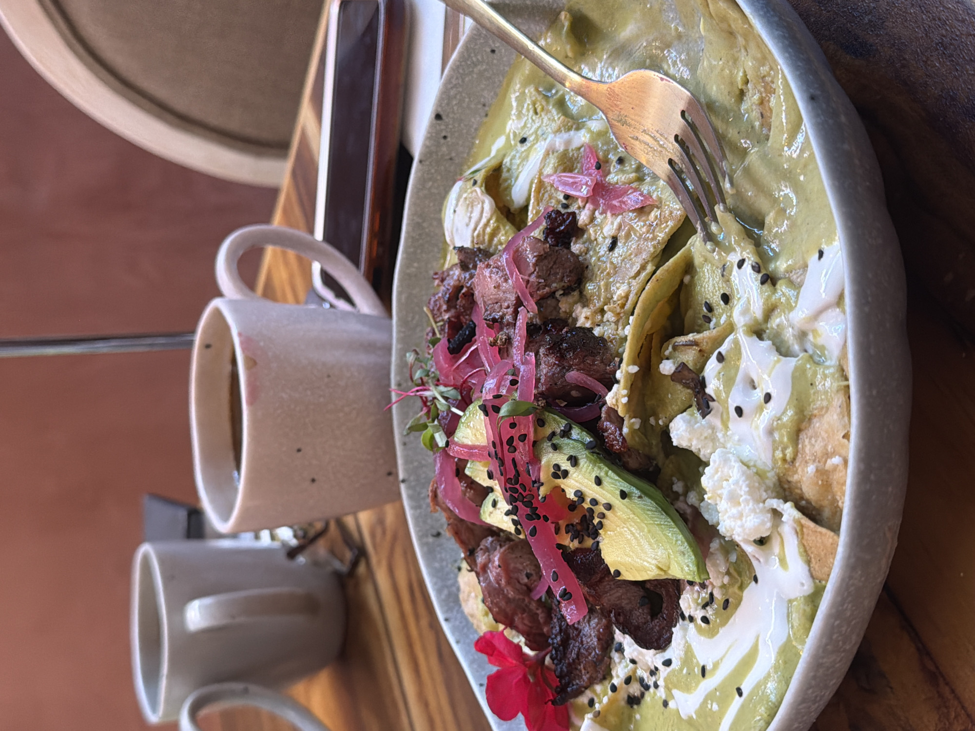 A close-up, high-angle shot captures a partially eaten plate of what appears to be Mexican food, specifically chilaquiles, along with two coffee mugs, likely in a restaurant setting. The chilaquiles are topped with meat, sliced avocado, pickled onions, cream, and a green sauce, possibly salsa verde. Black seeds are sprinkled across the dish. A fork is partially submerged in the food, suggesting it is in the process of being eaten.
Beside the plate are two ceramic coffee mugs, one positioned higher than the other. The higher mug is partially full, indicating it has been used.
The table is made of wood and positioned near a wall that appears to be peach or salmon-colored.
The scene is located in the city of Unknown, Unknown.