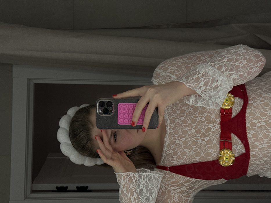 A woman, reflected in a mirror, holds a phone with a pink pop-it grip covering her face. She wears a white, lace, long-sleeved dress with a red, ornamented harness. Her hair is pulled back with a white, frilly headband. The background consists of a room with a white wall and window frame.