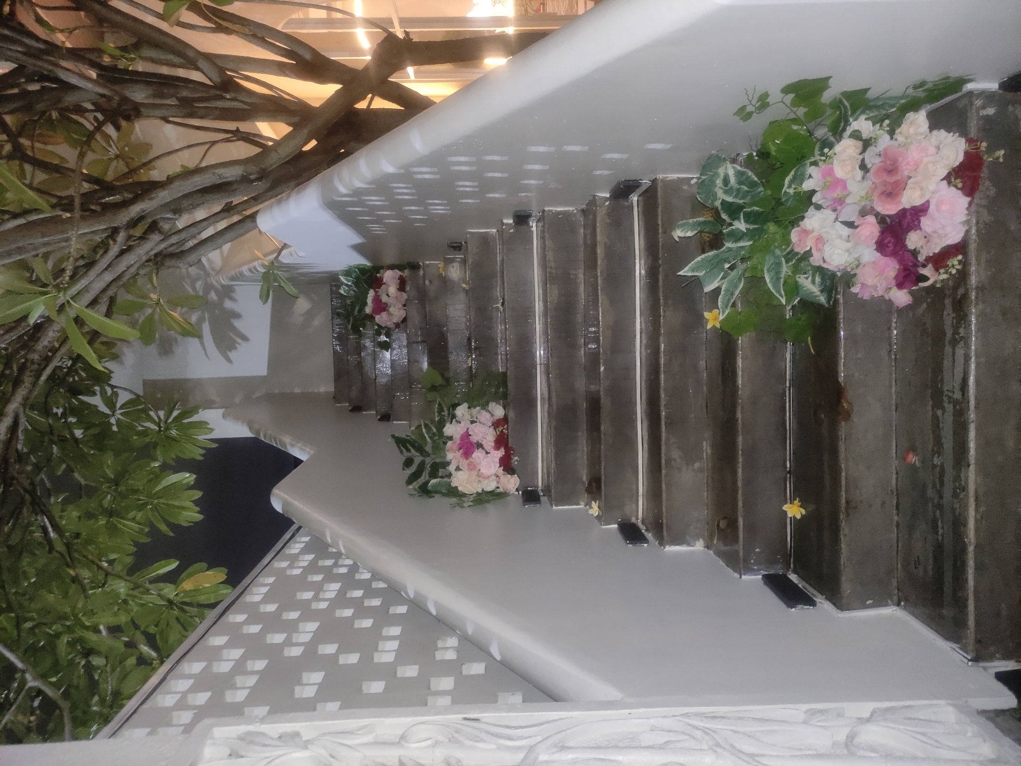 A view of a staircase decorated with floral arrangements, potentially for an event. The stairs are light gray, and the floral decorations include pink, white, and red flowers with green foliage. Some individual yellow flowers appear on the steps. The setting seems to be outdoors, with trees and foliage visible on the sides. The architecture has white details, suggesting a modern or contemporary style. The location is potentially in East Jakarta, Indonesia.