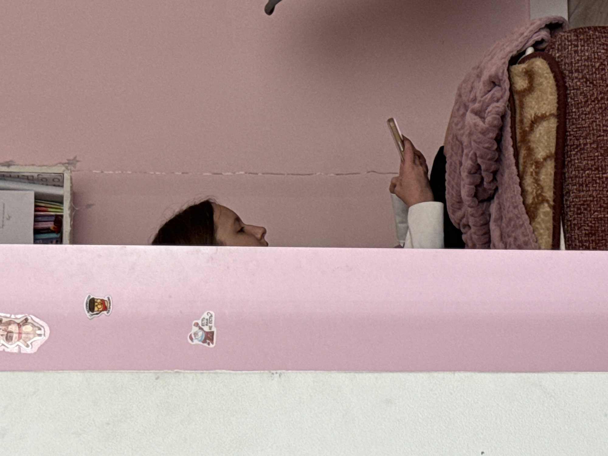 A person is lying down, looking at a phone. The person has dark hair and is wearing a white sleeve. There is a blanket to the right of the person. The background is pink. There are stickers on the pink surface in front of the person. The location is Unknown, Unknown.