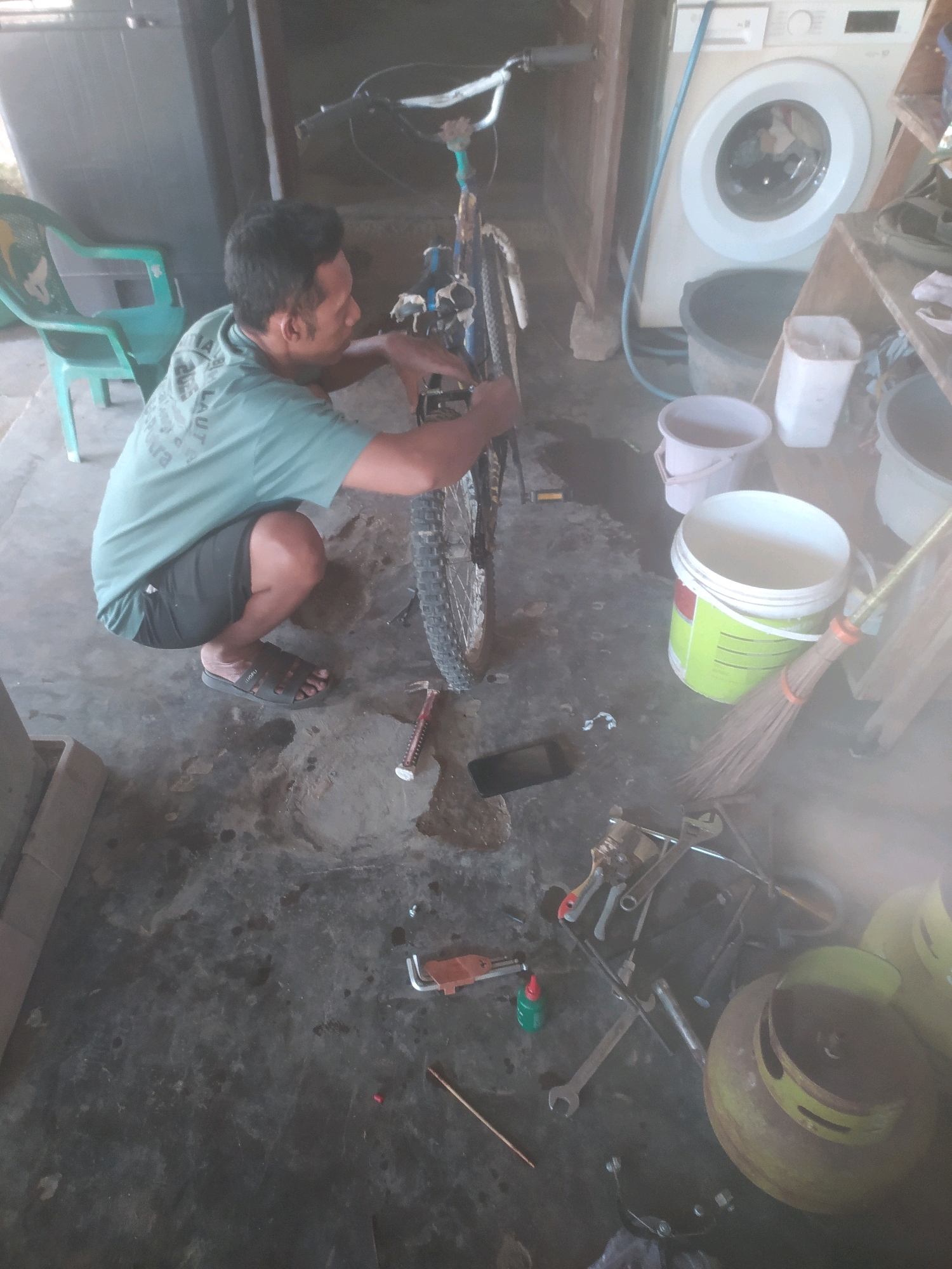 The image depicts an outdoor scene in Karanggayam, Indonesia, where a man is repairing a bicycle. The man, wearing a teal t-shirt and black shorts, is crouched down, actively working on the bicycle. Tools, including wrenches, are scattered around him on the concrete ground, indicating a repair in progress. A teal plastic chair is visible to the left, and a washing machine and various buckets are present in the background, suggesting a cluttered or utilitarian outdoor area. The overall environment appears somewhat unkempt, with water stains and debris on the ground.