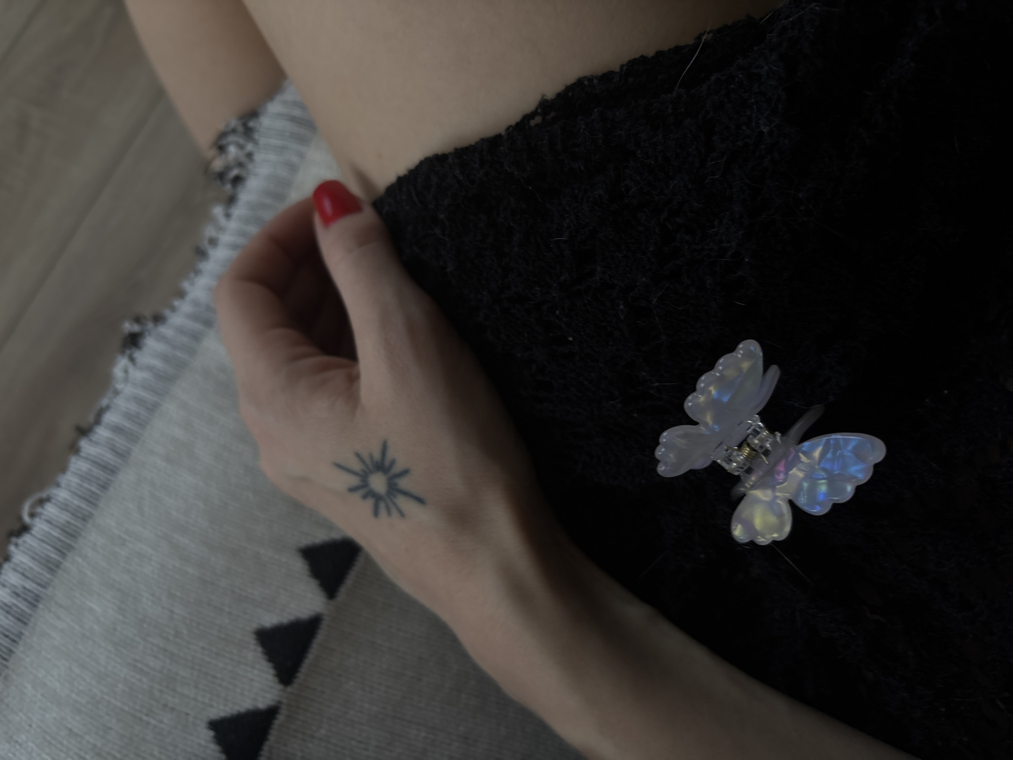 The image shows a close-up view of a person's hand with a sun tattoo on the back of it, resting on a dark, textured fabric. A small, iridescent butterfly-shaped hair clip is also visible on the fabric. The person's fingernails are painted red. The background consists of a grey, patterned cushion and a wooden floor. The image does not contain any information that can be used to determine its location within the city of Zheleznogorsk, Russia.
