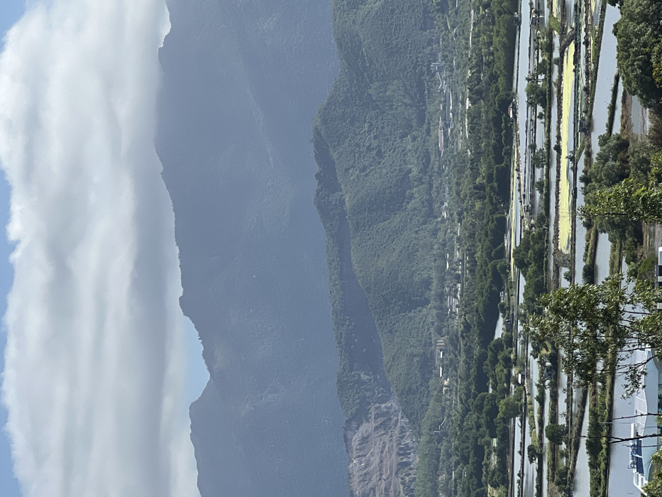 The landscape showcases a series of mountains, some shrouded in mist or clouds. Lush, green forests cascade down the slopes towards a valley where a small settlement is visible. Adjacent to the settlement are cultivated fields, possibly rice paddies, arranged in a grid-like pattern with water reflecting the sky. Trees are interspersed among the fields, adding to the area's verdant appearance.