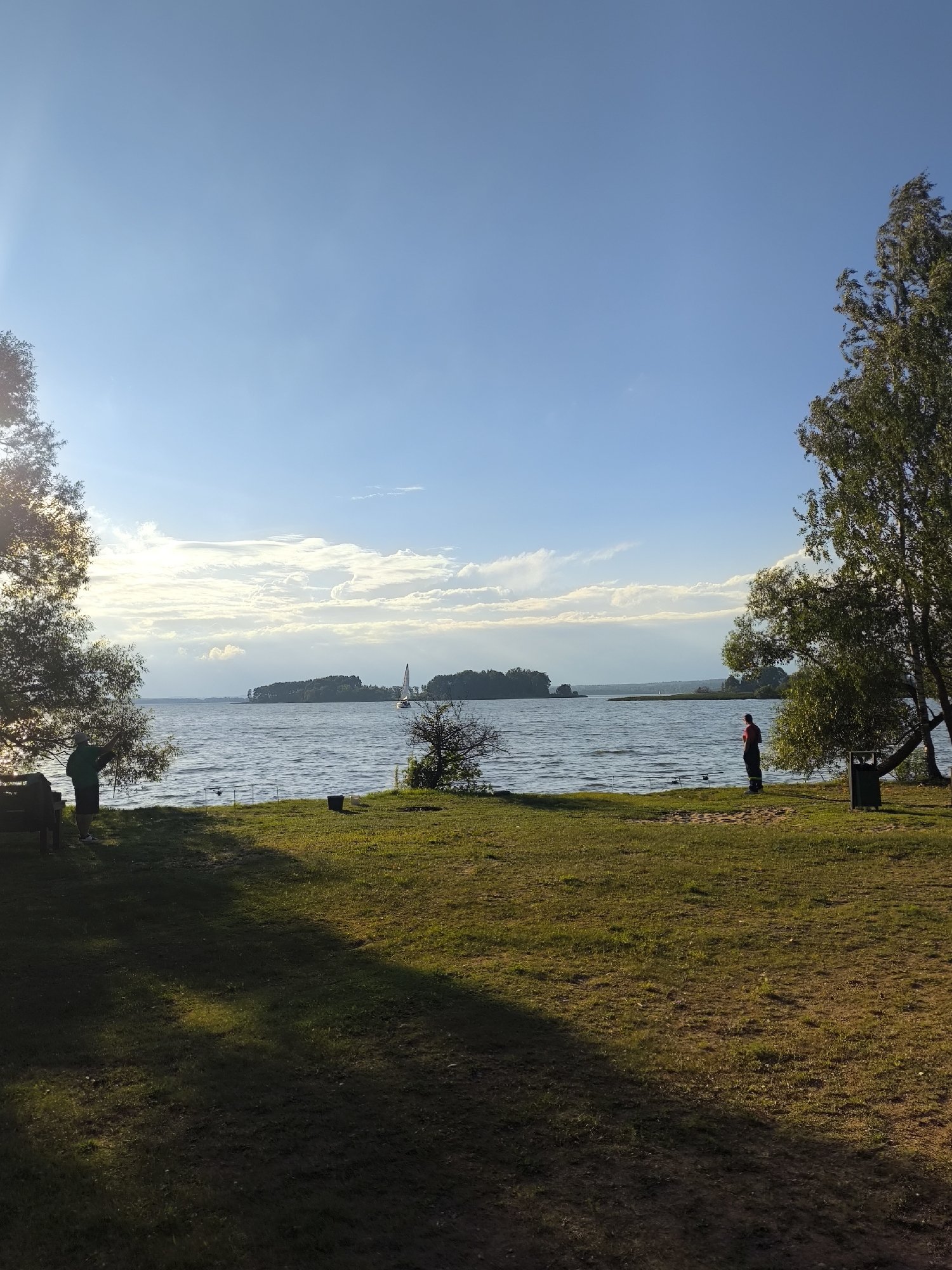 The image depicts a serene waterside scene under a bright, partially cloudy sky, likely in the vicinity of Ждановіцкі сельскі Савет, Belarus. The foreground features a grassy area with dappled sunlight and shadow, indicating late afternoon or early morning. A large tree with dense foliage is on the left, casting a long shadow. A dark-colored bench is visible near its base.
On the grassy bank bordering the water, two individuals are present. On the left, a person wearing a green shirt and dark shorts stands near the tree, facing towards the water. On the right, another person, dressed in darker clothing with a red accent on their upper body, stands further down the bank, also facing the water. Several fishing rods are set up on the bank between the two individuals, suggesting a fishing activity. A dark bucket is placed near the fishing rods.
The middle ground is dominated by a wide expanse of rippling water, likely a lake or a large river. In the distance, several tree-lined islands or peninsulas are visible. A white sailboat with its sails up is navigating the water. The horizon is marked by a line of trees and land. The sky is a clear blue with scattered white and gray clouds, illuminated by the sun which is positioned high and to the left, creating a bright haze. The overall atmosphere is calm and tranquil.