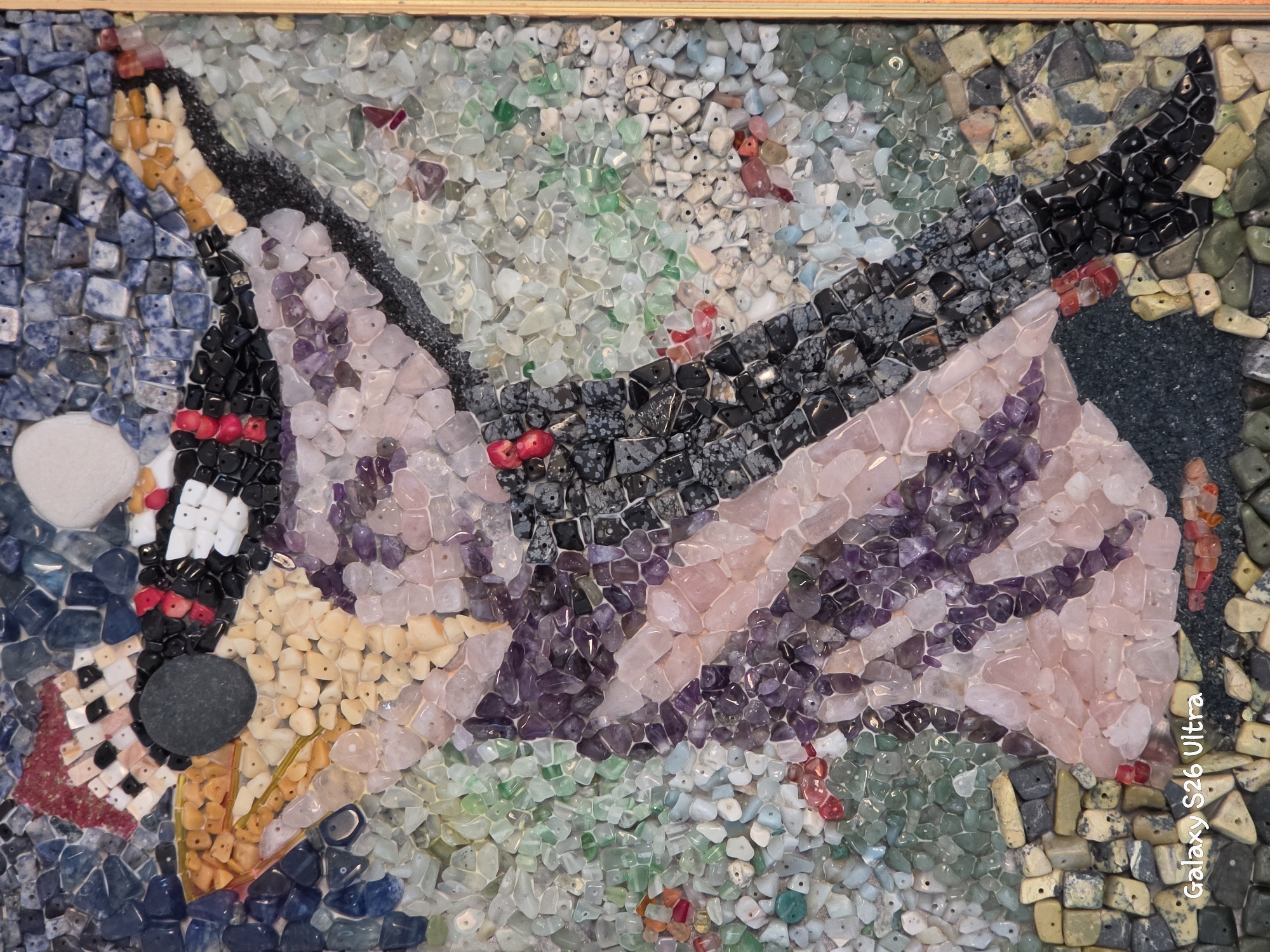 This is a close-up, full-shot, eye-level, indoor photograph of a mosaic artwork. The mosaic depicts a stylized human figure, possibly a dancer, rendered in various small, colorful stones. The figure is primarily composed of pink, purple, and black stones, with accents of red and orange. The background is a mosaic of green, blue, and white stones, giving the impression of water or a natural landscape. The mosaic is framed by a light-colored wooden border. There are no people or specific city locations identifiable within the image. The bottom left corner of the image contains a "Galaxy S26 Ultra" watermark.