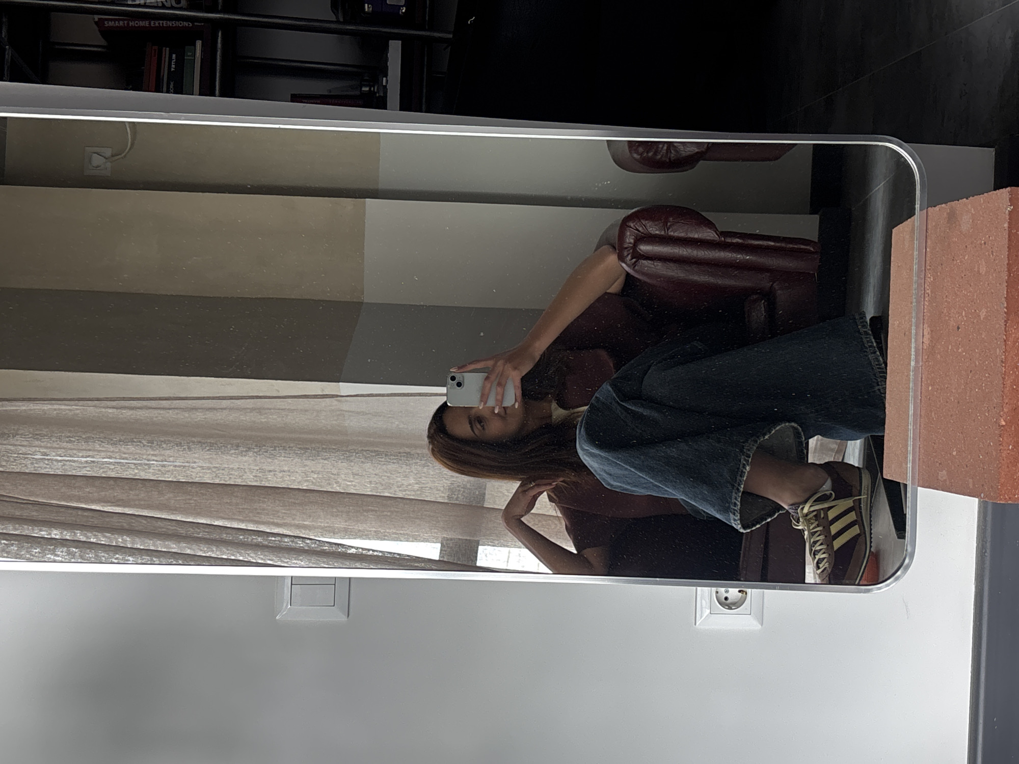 The image is a reflection in a mirror showing a woman sitting in a red leather armchair, holding a smartphone, presumably taking a selfie. The setting appears to be indoors. Behind the woman are curtains, and on the wall are electrical outlets and possibly a thermostat. In the background, a bookshelf is visible. The location is difficult to discern from the image but is stated to be Yerevan, Armenia.