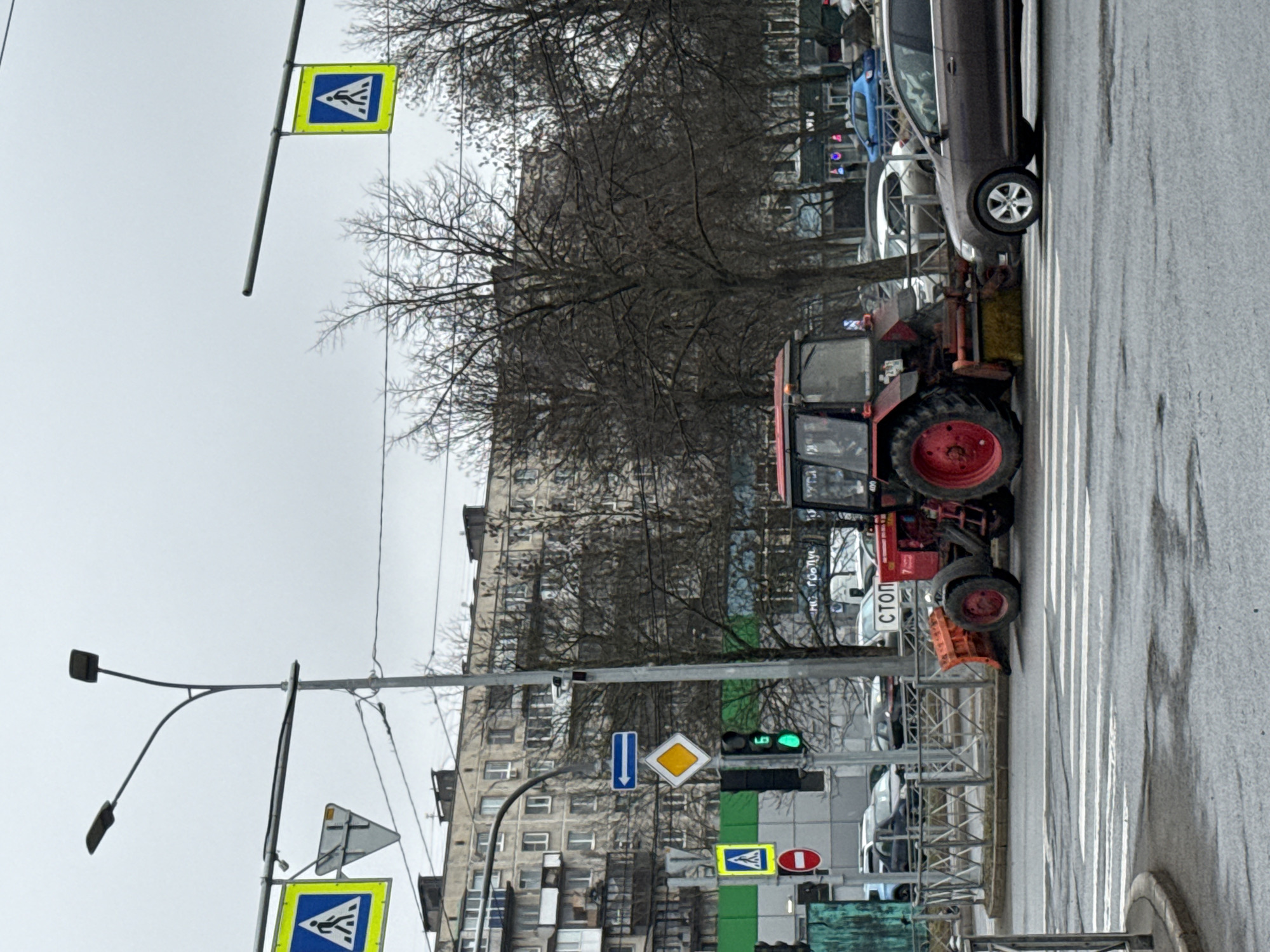 The image depicts a street scene in Saint Petersburg, Russia. A red tractor with a snow plow attachment is situated on a crosswalk next to a dark-colored car. Bare trees line the street, and a multi-story building stands in the background. Several road signs are visible, including pedestrian crossing signs, a stop sign (СТОП), and other traffic signs. A traffic light is also present, displaying a green light. The weather appears overcast. The road surface is wet, suggesting recent precipitation.