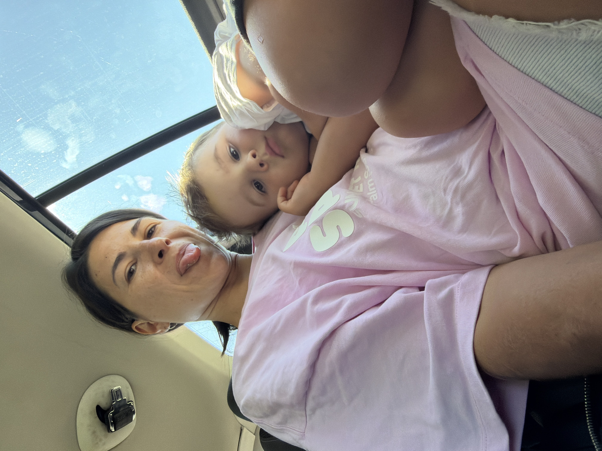This is a low-angle, close-up selfie taken inside a vehicle, likely a car, as indicated by the presence of a ceiling-mounted latch on the left and a sunroof with a view of the sky above. The image features two individuals: a woman and a young child. The woman is in the foreground, to the left, and is sticking her tongue out at the camera. She has dark hair pulled back and is wearing a light pink oversized t-shirt with a white graphic design. To her right, nestled against her, is a young child, likely a boy, resting his head on her chest. The child has light brown hair and is looking towards the camera with a slightly weary expression. He is wearing a white t-shirt. The overall setting appears to be daytime, with sunlight streaming through the sunroof, creating bright highlights and a slightly hazy atmosphere. The interior of the vehicle is dimly lit, with the exception of the area illuminated by the sunroof. The background is dominated by the sky, which appears blue with some scattered clouds. The scene conveys a sense of intimacy and a candid moment between the two subjects. There is no indication that this image was taken in the city of Kruševica, Montenegro.