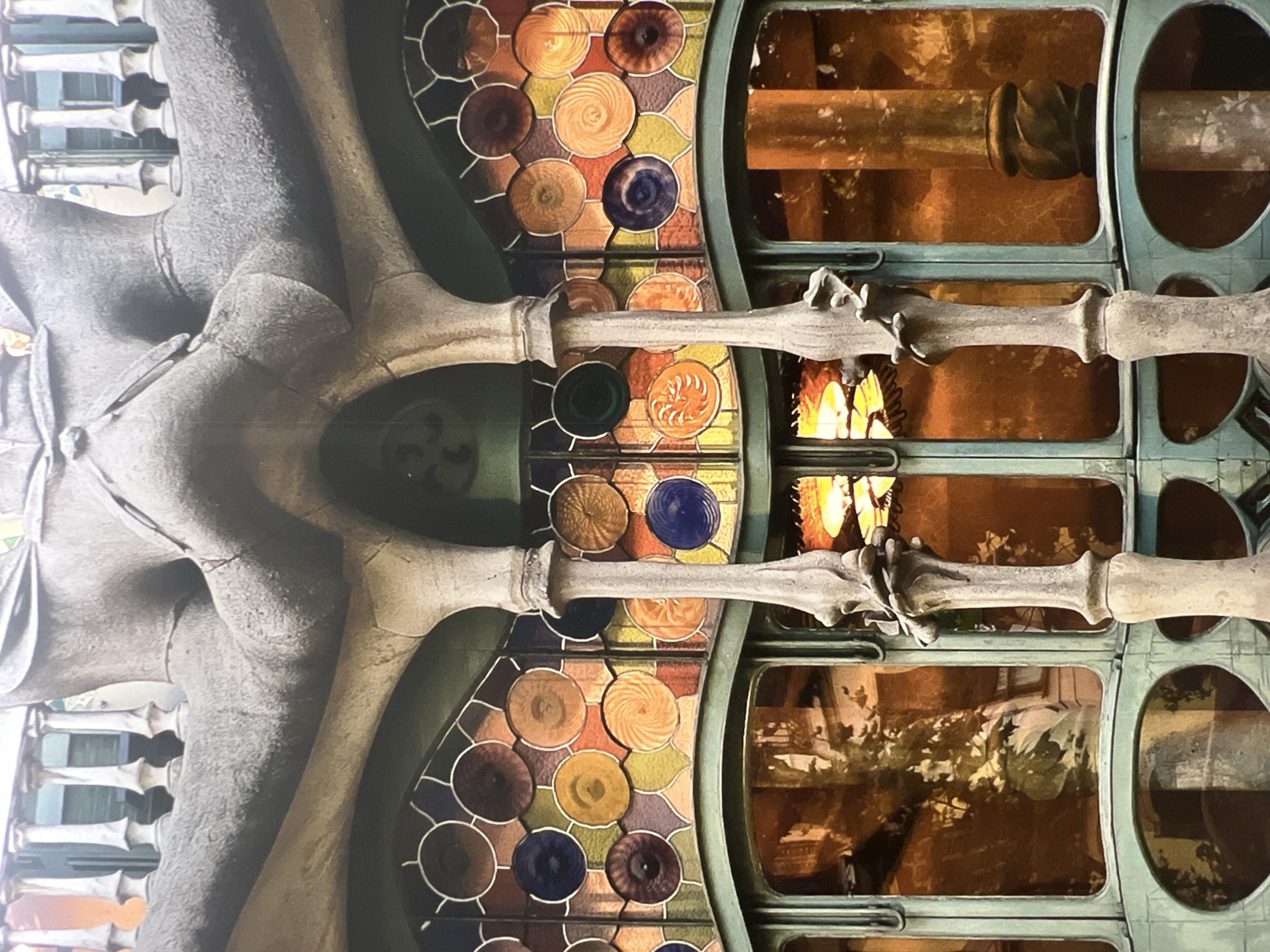 This is a photograph of the facade of Casa Batlló, a building designed by Antoni Gaudí, located on Passeig de Gràcia in Barcelona, Spain. The image focuses on the upper portion of the building, specifically a large, organic-shaped balcony with a stone railing and decorative columns. Below the balcony is a vibrant stained-glass window composed of numerous circular panes in various colors, including shades of orange, yellow, blue, and purple. The stained glass has a mosaic-like appearance with intricate patterns within each circle. The windows below the stained glass are made of metal frames and large panes of glass, reflecting warm, amber light and the silhouettes of trees or plants. Two tall, slender, stone columns with elaborate, sculptural bases and tops support the balcony, framing the central section of the stained-glass window. The overall aesthetic is characteristic of Gaudí's modernist style, with flowing lines, naturalistic forms, and a rich use of color and texture.