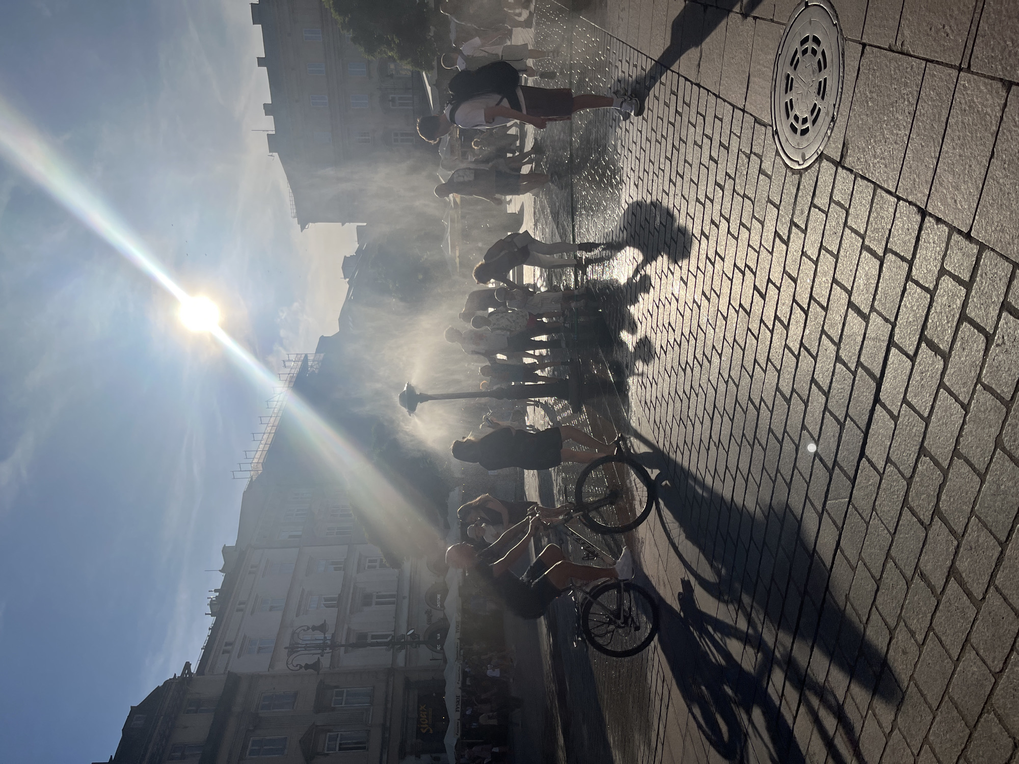 The image depicts a sun-drenched urban square in Krakow, Poland, likely Rynek Główny (Main Market Square), given the architectural style of the buildings and the presence of what appears to be a fountain or water feature. The sun is bright, casting long shadows across the cobblestone pavement. Several people are visible, some riding bicycles, others standing or walking, many interacting with a water spray emanating from a central point, suggesting it's a hot day and people are cooling off. The foreground shows a close-up of the cobblestone ground and the shadow of a bicycle. In the background, traditional European buildings with multiple stories line the square, some with awnings and outdoor seating areas. The overall atmosphere is lively and summery.