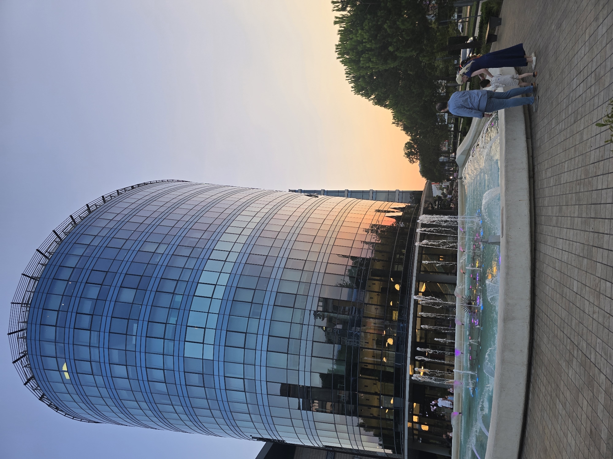 In the city of Podgorica, Montenegro, a modern cylindrical glass building stands tall against a gradient sky transitioning from blue to orange, indicative of dusk. The building's facade reflects the warm hues of the sunset, along with other structures and trees. In the foreground, a family of three, consisting of a man in a blue shirt and jeans, a woman in a long dark blue dress, and a small child in a light-colored outfit, are gathered by a large, multi-tiered fountain. The fountain features numerous water jets, some illuminated with pink and blue lights, creating a dynamic and colorful display. The area around the fountain is paved with interlocking bricks. To the right of the fountain and building, a row of tall, green trees lines a street with a few cars visible. The overall scene captures a tranquil urban evening ambiance.