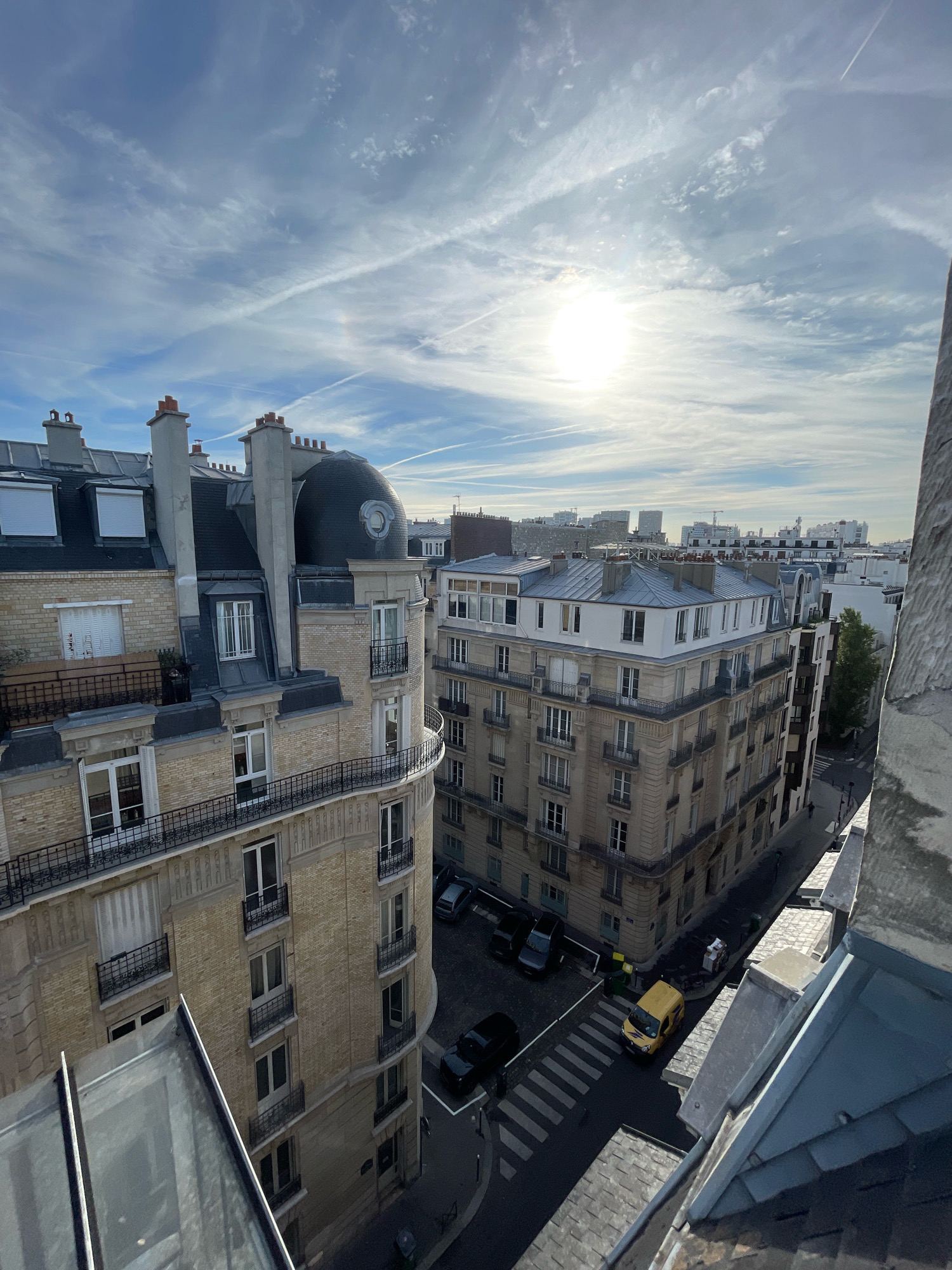 An aerial, high-angle, medium shot captures an overcast sky with a bright, diffused sun partially obscured by clouds, producing a halo effect. The sky is streaked with contrails from aircraft. Below the sky, a dense urban cityscape of Parisian buildings is visible. The buildings are primarily constructed of light-colored brick and stone, featuring mansard roofs with multiple chimneys and dormer windows. Several balconies with black iron railings adorn the facades. In the street below, a yellow delivery van is parked on a crosswalk, and several cars are parked along the side of the road. The scene suggests an urban environment in Paris, France, likely during daylight hours.