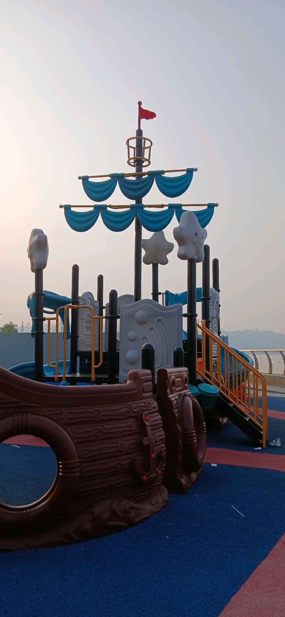 The image depicts a pirate ship-themed playground structure situated on a blue and red rubberized surface. The playground features a central mast with a red flag at its apex and several blue, wave-like decorative elements resembling sails. White, star-shaped decorative elements are also attached to the structure. Various slides and climbing structures are visible, some with yellow railings. In the foreground, a large, brown, textured piece of the ship, possibly a prow or hull section, is prominent. The background shows a hazy sky and a body of water, suggesting an outdoor location. There are no people visible in the image. The overall scene appears to be a public recreational area.