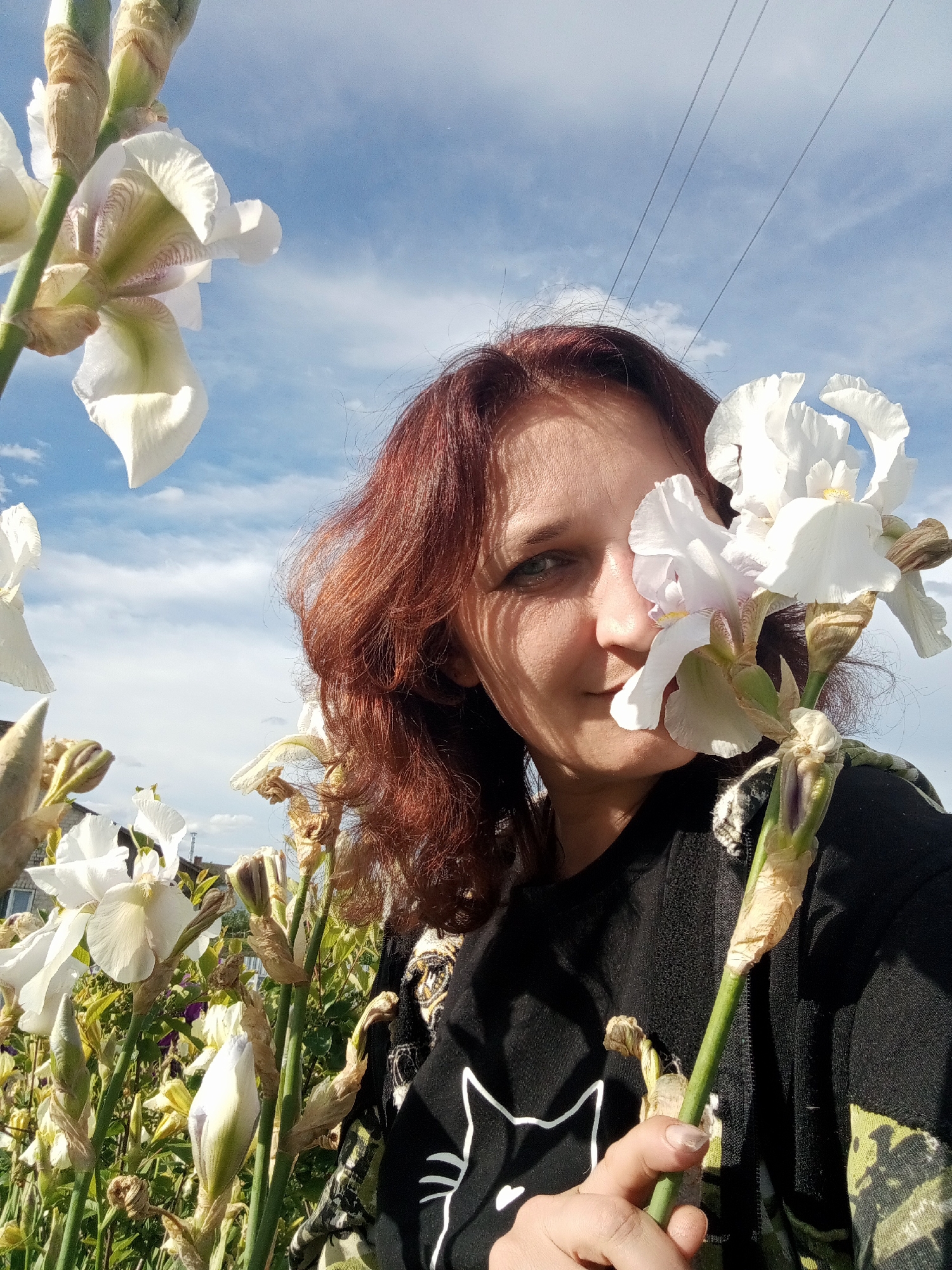 A woman with auburn hair poses in a garden filled with white irises under a blue sky with scattered clouds. She holds a white iris flower close to her face, partially obscuring one eye. She wears a black shirt with a white cat graphic. The background includes power lines, a glimpse of a building, and other irises in various stages of bloom. The scene is bathed in sunlight. The location is Babyn, Ukraine.