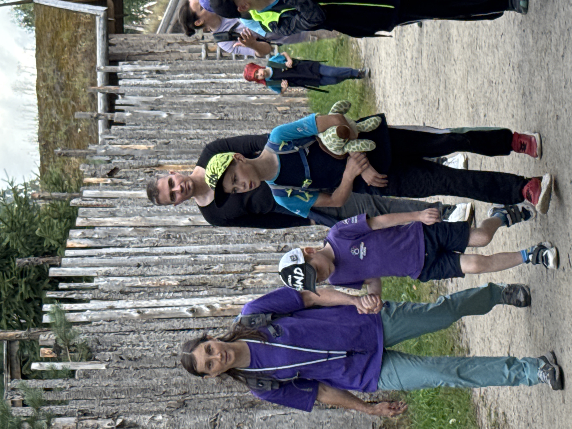 In Khankanmyaki, Russia, a group of people are walking along a dirt path next to a rustic wooden fence. The fence is made of vertical, rough-hewn logs, with some logs showing evidence of charring. Beyond the fence, a building with a thatched roof can be seen. In the foreground, a woman in a purple shirt and light blue pants holds hands with a young boy wearing a purple shirt and dark shorts. The boy is wearing a black and white baseball cap. Beside them, a teenage boy in a blue shirt and black pants walks, carrying a stuffed turtle and wearing a lime green baseball cap. Behind them, other individuals are visible walking along the path, including a man in a black shirt and a woman in a light purple shirt. The overall scene suggests an outdoor excursion or a visit to a historical or cultural site.