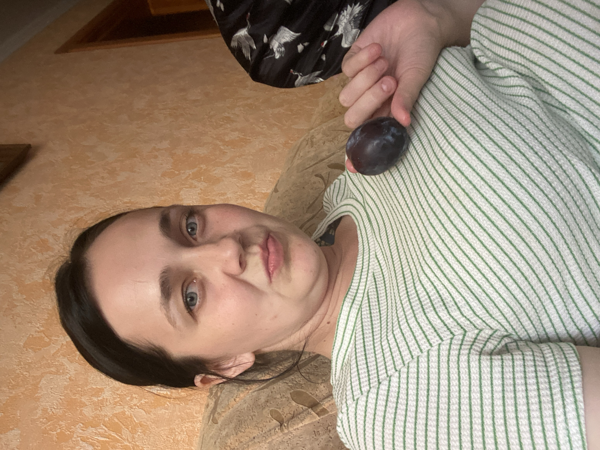 A Caucasian woman is shown from the chest up, sitting in a light brown textured armchair against an orange textured wall. She has dark brown hair pulled back, light blue eyes, and is making a puckered face. She is wearing a green and white horizontally striped shirt. Her right hand is raised, and she is holding a dark purple plum. The background shows a portion of a doorway and a black garment with a white bird pattern. The lighting appears to be indoor and warm.