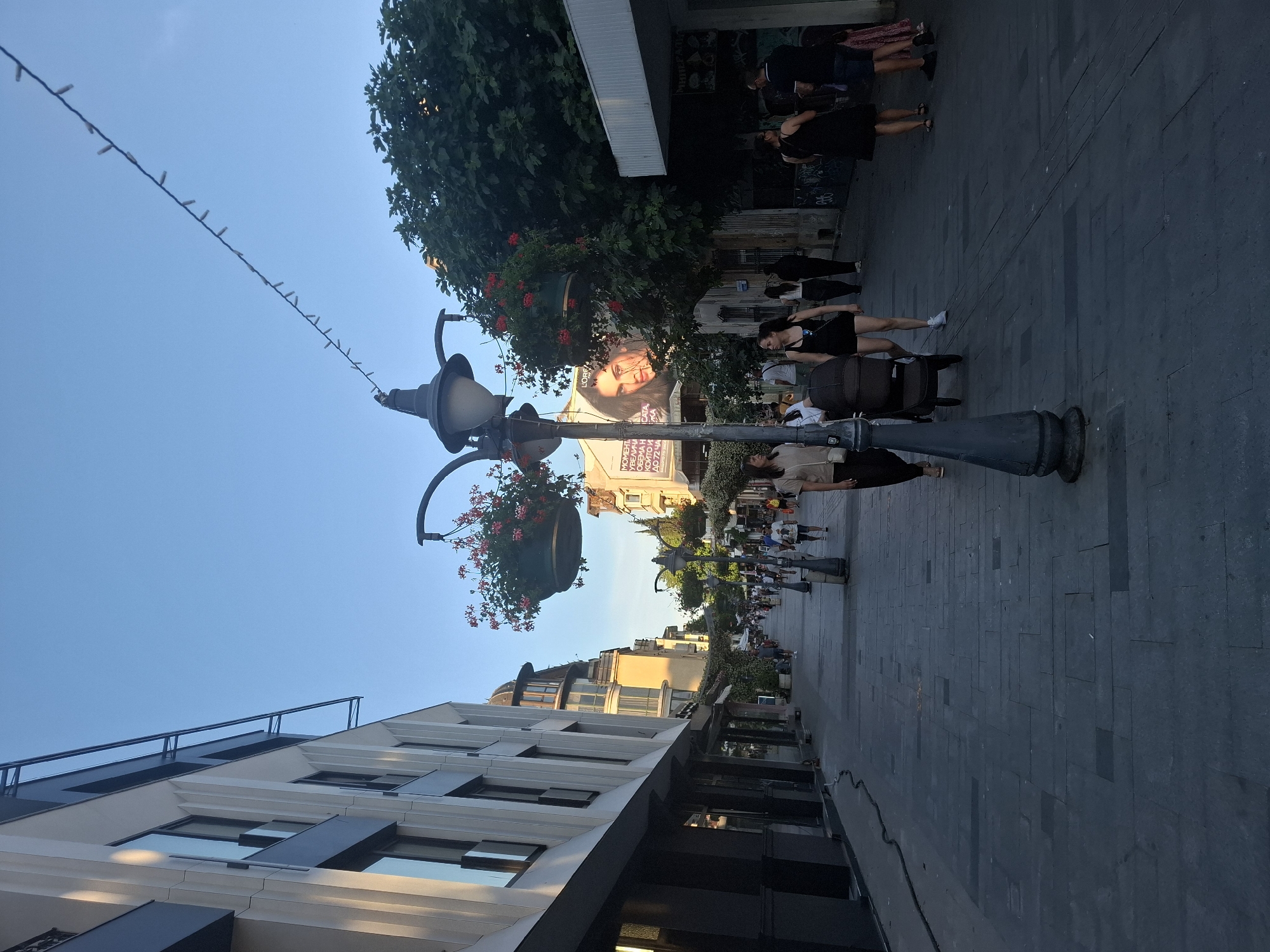 The image shows a pedestrian street in an urban environment. Modern buildings with large windows line the left side of the street, while older buildings and trees are visible on the right. String lights are suspended across the street above the pedestrian walkway. Several people are walking on the street, some are pushing strollers, and others are standing and talking. There are also hanging flower baskets on lampposts. In the background, a large billboard with a woman's face is visible. The sky is clear and blue.