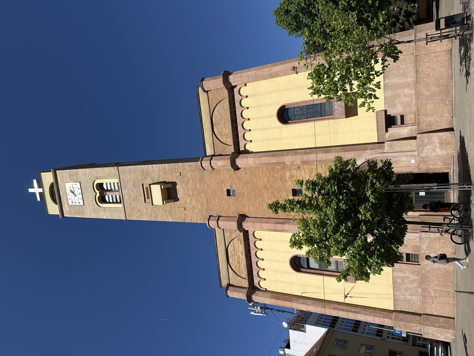 This image captures a vibrant, sunny day in Zagreb, Croatia, showcasing a distinctive church building with a tall bell tower. The church features light yellow plastered walls accented with reddish-brown stone or brick decorative elements, including arched cornices and pilasters.
The prominent bell tower on the left is primarily constructed of reddish-brown brick and culminates in a white cross. A clock face on the tower, clearly visible, indicates the time is approximately 10:25. Below the clock, arched openings suggest a belfry. The main body of the church includes windows, some appearing to be stained glass.
The sky is a deep, clear blue, signifying bright and pleasant weather. A faint airplane contrail is visible in the upper sky. Several trees with lush green foliage, suggesting spring or summer, frame parts of the church, with one particularly prominent tree in the mid-ground partially obscuring the building. Bright sunlight casts distinct shadows, indicating late morning or early afternoon.
On the paved sidewalk in the foreground, two people are visible. One individual, dressed in a light-colored jacket and pants, walks away from the church. Another person is partially seen behind a tree, near a bicycle parked at a rack. Other urban structures, including residential or commercial buildings and modern communication antennas, are visible in the lower sections of the image, positioning the church within a bustling city environment. A blue parking sign with a white "P" is also discernible on the street level.