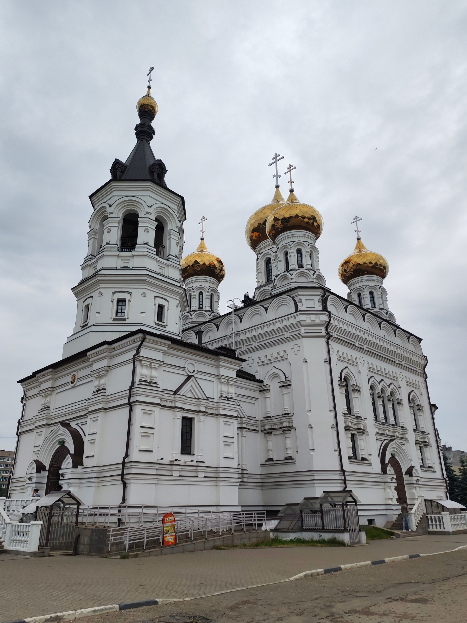 This is a medium-angle, low-fidelity, outdoor photograph of the Ascension Cathedral in Tver, Russia, taken from street level on an overcast day. The cathedral, a large, white, multi-domed Orthodox church with gold-colored domes and silver crosses, dominates the frame. The main structure features multiple levels with arched windows and decorative white plasterwork. A tall bell tower on the left has a black roof and a golden dome. In the foreground, a paved sidewalk and street area are visible, with a white railing and a ramp leading up to an entrance of the church. A red and yellow informational sign is placed near the ramp. A few people are visible in the scene: one standing near the entrance on the right, another partially visible in an entrance on the left, and some on a rooftop or elevated platform on the church itself. The sky is cloudy and light grey. The image captures the architectural grandeur of the cathedral against a somber sky.