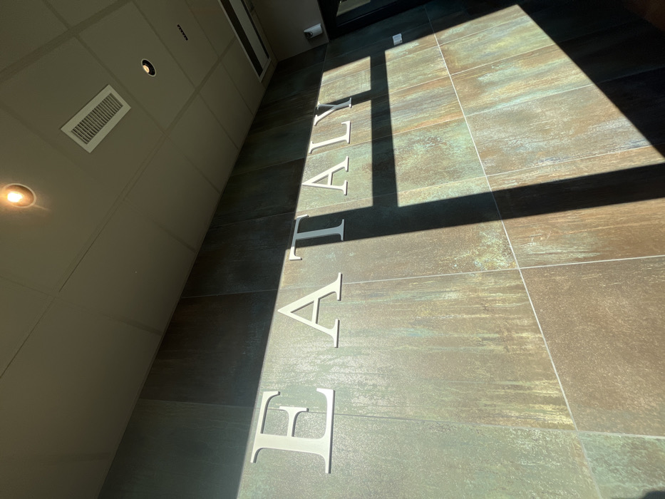 The image captures an interior view, likely a hallway or entrance, characterized by a contemporary and clean design. The perspective is an upward diagonal shot, with a tiled floor occupying the right and central portion, and a light-colored wall or ceiling dominating the left side.
Dominating the floor are large, three-dimensional silver or white letters spelling "EATALY". These letters are evenly spaced along the floor, running parallel to the wall on the left. The floor itself is composed of large, rectangular tiles with a textured, variegated appearance, featuring tones of brown, green, and grey, giving it a somewhat distressed or industrial look.
Strong, natural sunlight streams into the space from the right, casting sharp, elongated shadows across the floor and partially onto the letters and the adjacent wall. This indicates a bright, sunny day, likely morning or afternoon when the sun is at an angle. The contrasting interplay of bright light and deep shadows creates a dynamic visual effect.
On the left-hand wall/ceiling, which is a light neutral color (beige or light grey), there are several architectural elements. Two recessed circular lights are visible, one brightly illuminated in the bottom left, and another further up. A rectangular air vent or grille is also prominently featured on this surface. A dark doorway or frame is partially visible in the upper right corner, adding depth to the scene. No people are present in the image.
The visible text "EATALY" clearly indicates the location is associated with the Eataly brand, an Italian marketplace chain. Given the provided location context of Dorno, Italy, this image likely depicts an Eataly establishment in or around that area.