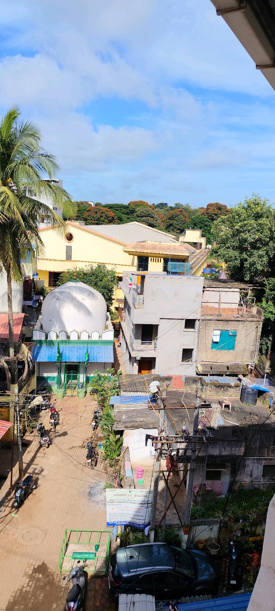 This is a high-angle, vertical shot of a street scene in Belagavi, India, likely taken from a rooftop or balcony. The sky above is blue with scattered white clouds. In the foreground, a large palm tree with green fronds is visible on the left. The street below is paved with interlocking bricks and has several parked motorcycles and scooters.
Dominating the mid-ground is a white domed structure, possibly a mosque, with green trim and two green flags. Adjacent to it is a yellow building with a large sloped roof. Several multi-story buildings, some appearing aged with exposed brickwork and tin roofs, are scattered throughout the scene. Power lines and electrical poles are prominent features, indicating the urban setting.
Vegetation is present, including trees with green foliage and some trees with orange flowers in the background. The scene depicts a typical day in a densely populated urban area, with evidence of daily life like vehicles and residential buildings. There are no visible people engaged in specific actions, but the presence of parked vehicles suggests ongoing activity. A black car is parked at the lower right of the image. A blue banner with text is hanging on a wall.