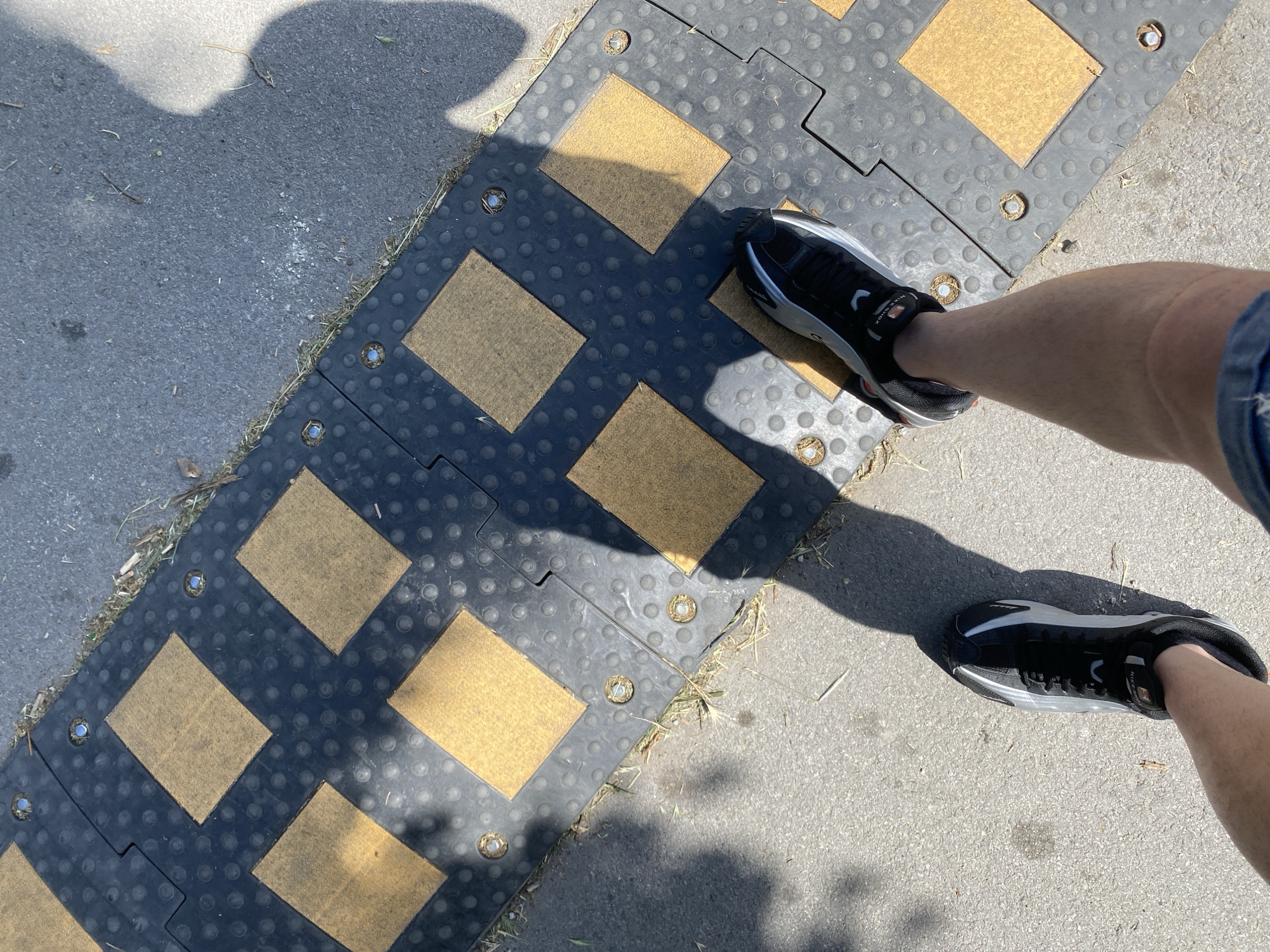 An overhead shot captures a person's legs and feet standing on asphalt next to a speed bump. The speed bump is made of black rubber with raised yellow squares and circular dimples. Metal fasteners secure the speed bump to the ground. A shadow of a person is cast across the speed bump and asphalt. The lower legs of the person are visible, with one foot wearing a black and white athletic shoe. The surrounding environment appears to be an outdoor urban setting, possibly in Varna, Bulgaria, given the context.