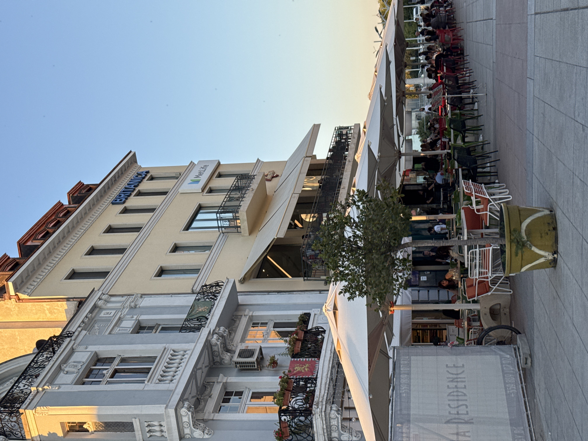 The image shows a street view in the city of Plovdiv, Bulgaria, on a sunny day. In the foreground, there is an outdoor seating area for a restaurant or cafe, with tables and chairs set up under large white umbrellas. Several people are seated at the tables, some appearing to be dining or socializing. A young tree in a large pot stands to the right of the seating area. Behind the seating area, there are multi-story buildings with shops and businesses on the ground floor. One building has a sign that reads "BRITANICA" in blue letters, and another has a "MetLife" logo. The architecture of the buildings suggests a historic European city. The sunlight casts long shadows, indicating it is likely late afternoon or early evening. A construction barrier with "TA RESIDENCE" written on it is visible to the left.