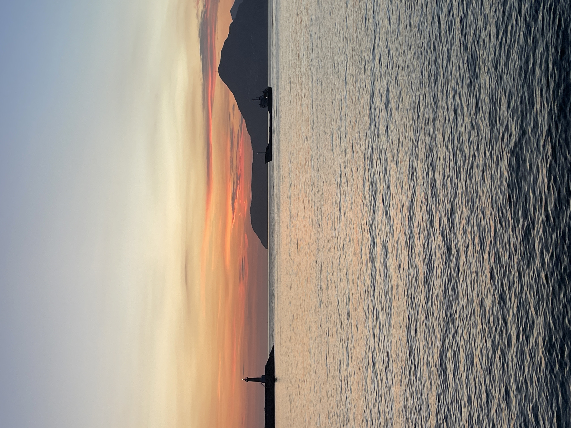 A serene seascape at dusk features a calm body of water reflecting the soft hues of the sky. In the distance, a lighthouse stands on a rocky outcrop to the left, its light illuminated. Further to the right, a cargo ship sails across the water. Behind the ship, a silhouetted mountain range borders the horizon. The sky displays a gradient of colors, from a pale blue at the top to warm oranges and pinks near the horizon. The image captures the tranquil atmosphere of the Bar coast in Montenegro.