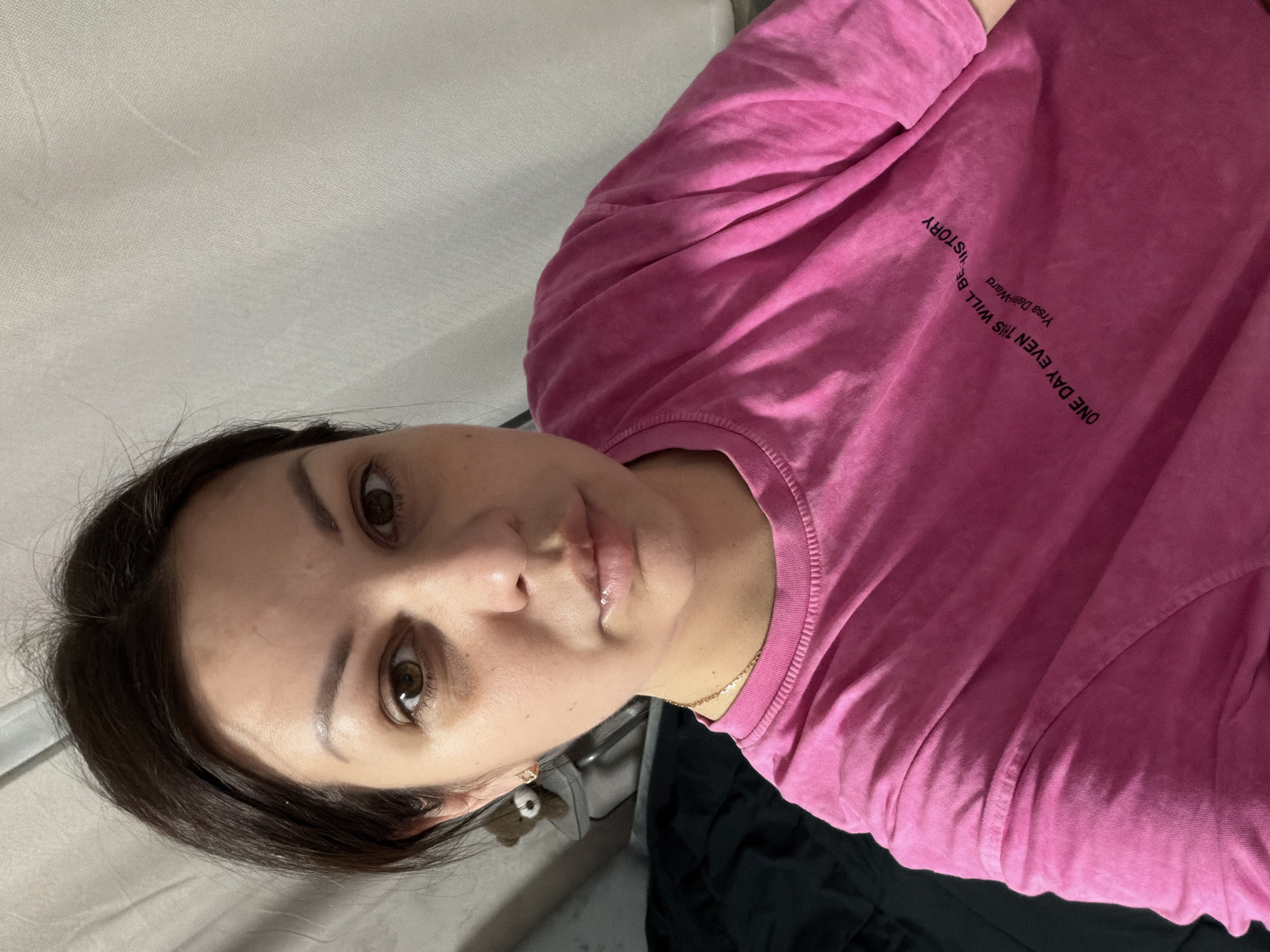 A woman is captured in a close-up, front-facing selfie, her gaze directed towards the camera. She has dark hair and brown eyes. She is wearing a pink t-shirt with text on it. The text on the t-shirt reads "ONE DAY EVEN THIS WILL BE HISTORY" and "Yrsa Daley-Ward". She is also wearing a gold necklace. The background consists of a textured white surface and a dark, possibly black, fabric, which could be a bed or blanket. A small teddy bear can be seen on the left side of the image. The lighting appears to be artificial, possibly from an indoor setting. There is no indication of the location being in Sochi, Russia.