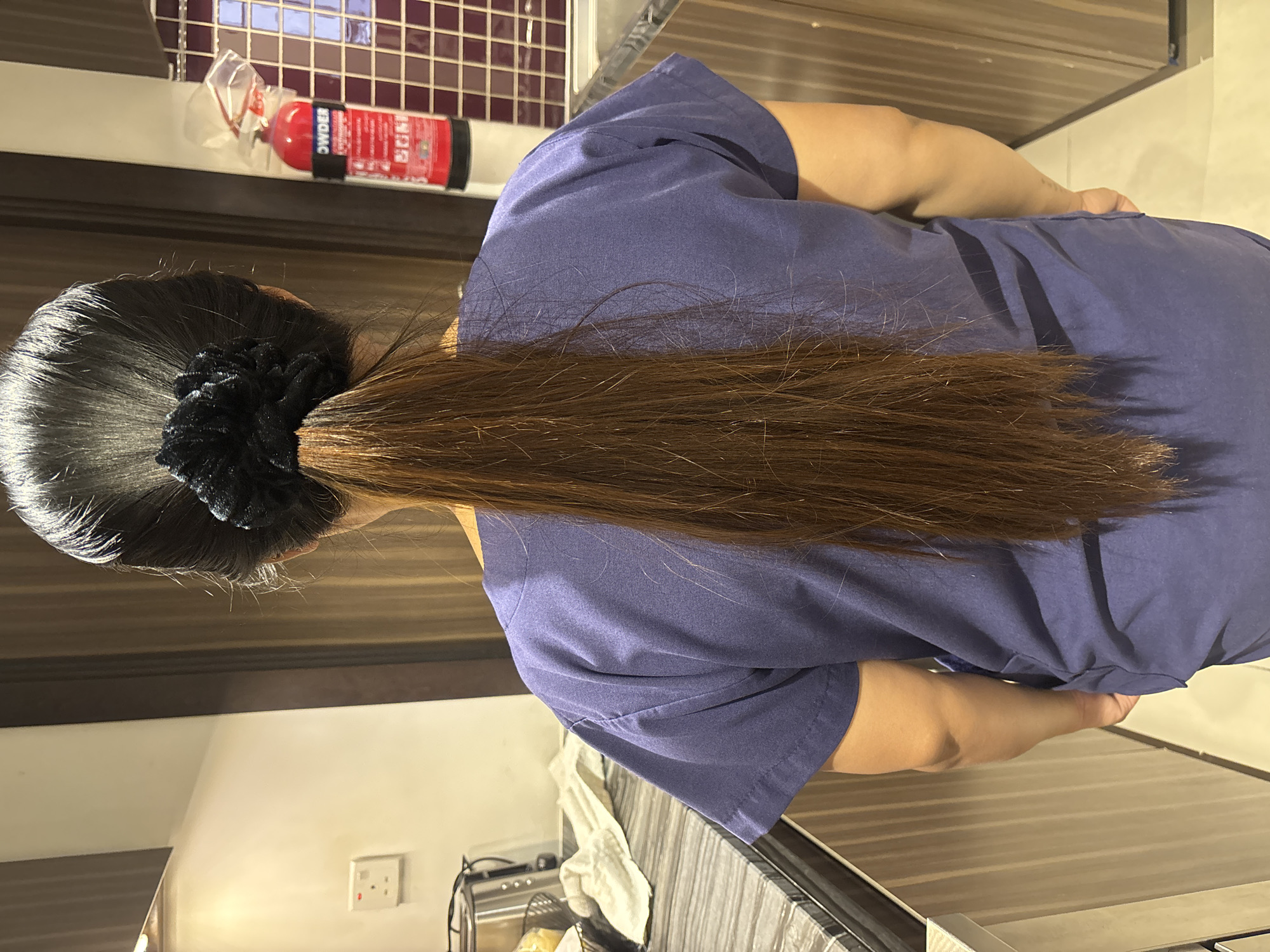 A rear view of a person's head and shoulders is captured in a medium shot, set against a domestic kitchen backdrop in Dubai, United Arab Emirates. The person's dark hair is pulled back into a high ponytail, secured with a black scrunchie. Their gaze is directed forward, away from the camera. The environment appears to be a kitchen, with wooden cabinets and a tiled backsplash visible. A red fire extinguisher is mounted on the wall to the right of the frame. The person is wearing a navy blue t-shirt.
