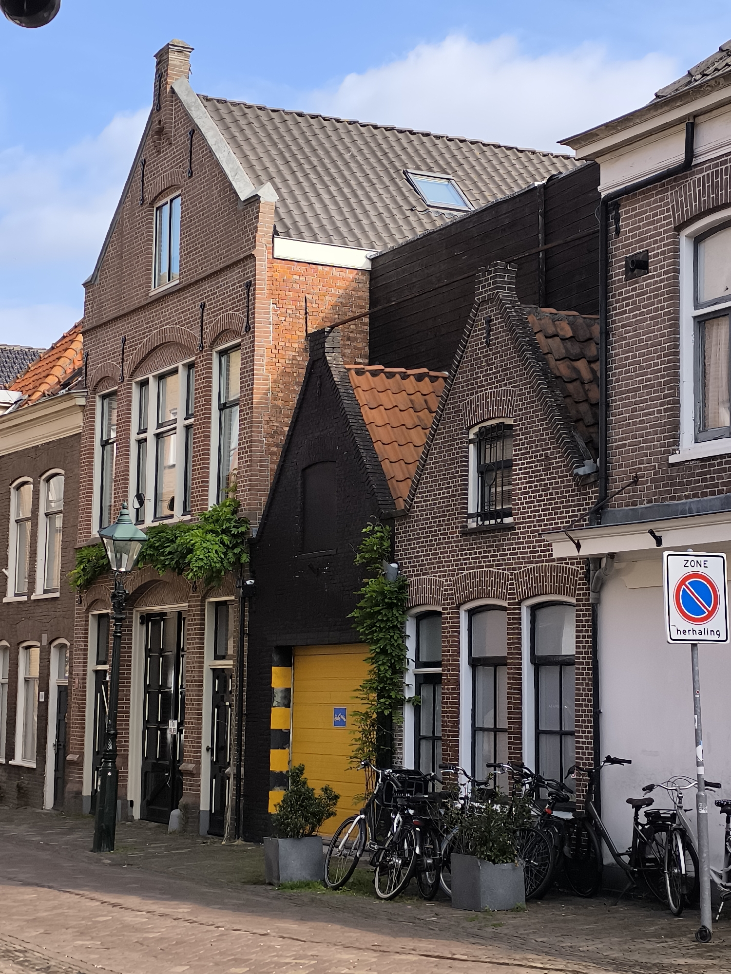 This is a daylight, eye-level, medium shot of a street scene in Alkmaar, Netherlands. The focus is on the facades of several brick buildings lining a cobblestone street.
The buildings are narrow, tall, and appear to be older architecture, with varying rooflines and materials. The building on the far left has a light-colored facade with multiple windows and an orange tiled roof. Next to it is a reddish-brown brick building with a steeply pitched, grey tiled roof and a prominent chimney. It features several windows with dark frames and decorative brick arches above them. A green metal lamppost stands in front of this building. To the right of that is a black facade building with a steeply pitched orange tiled roof. This building has a bright yellow garage door. Further to the right, another reddish-brown brick building is visible, with large windows and a white awning.
In front of the buildings, several bicycles are parked along the sidewalk. A potted plant sits near the yellow garage door, and another is placed near the row of parked bicycles.
A traffic sign is visible on the right side of the image. It is a white rectangular sign with a red border and a blue circle with a red diagonal bar, indicating a parking restriction. The word "ZONE" is at the top, and "herhaling" is at the bottom.
The sky is partly cloudy with a pale blue hue. The light suggests it is either morning or late afternoon, casting shadows on the street. There are no people visible in the scene. The overall impression is a quiet, historic street in a Dutch city.