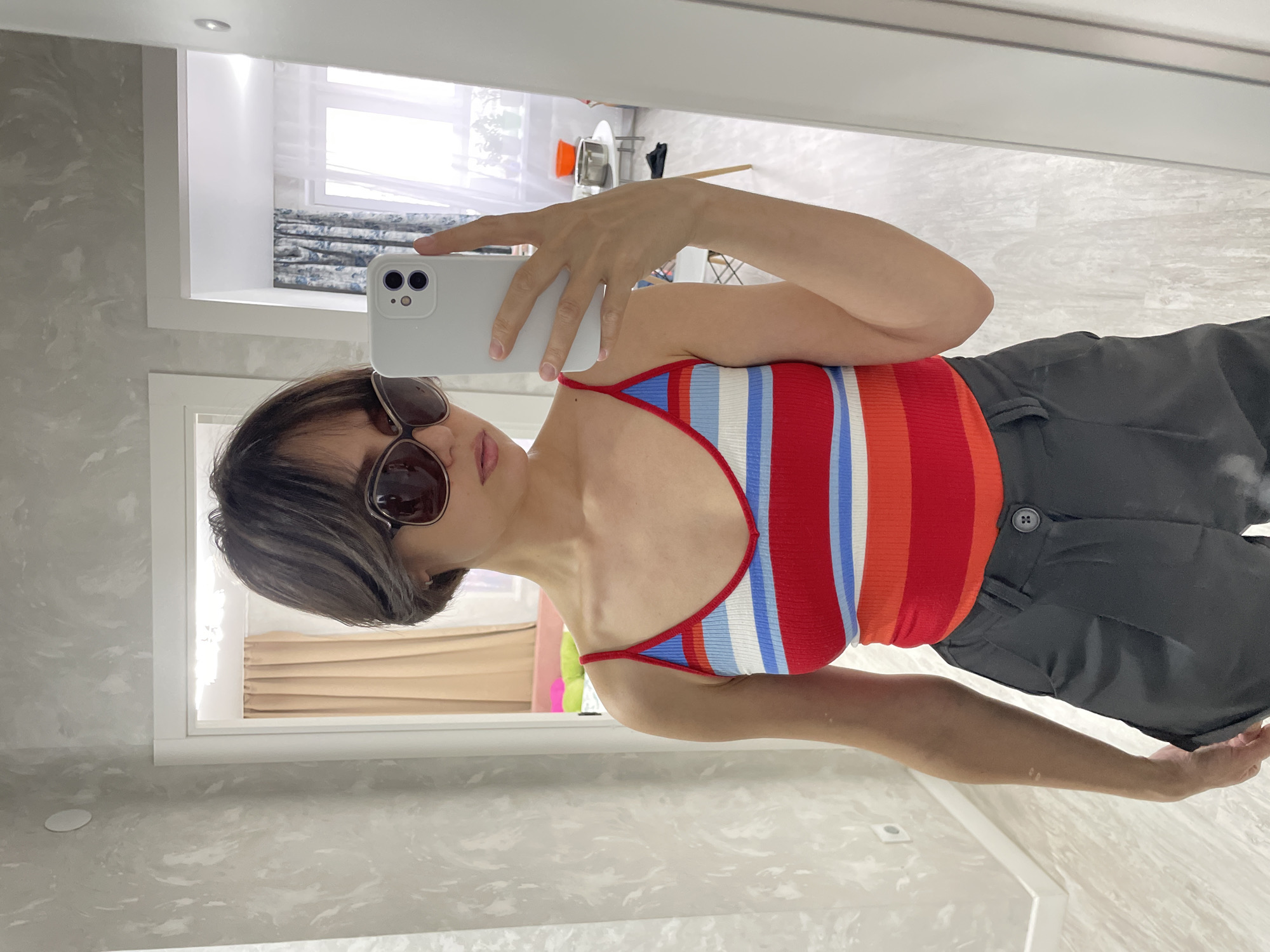 A woman is taking a selfie in a room. She is wearing sunglasses, a striped top with red, white, and blue, and dark grey pants. She is holding a white smartphone in her right hand.
In the background, there is a window with white frames and curtains. The room appears to be a bathroom, judging by the marble-patterned wall. The location is not identifiable.