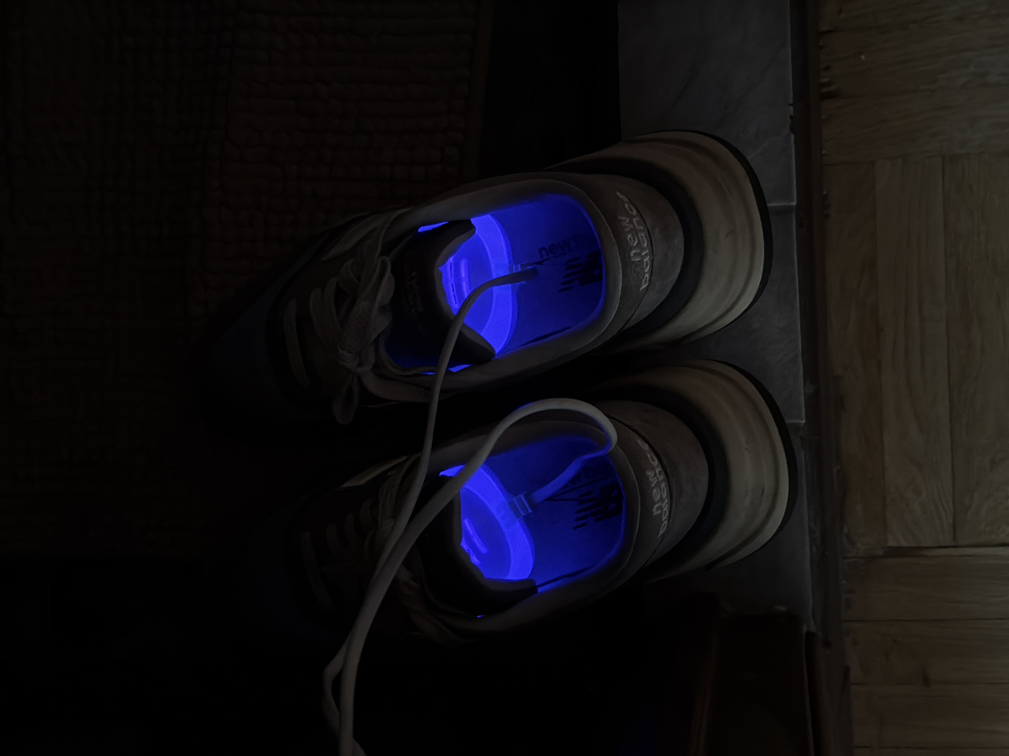Two New Balance athletic shoes are depicted in a dimly lit indoor setting. The most prominent feature is the bright, glowing blue light emanating from the interior of each shoe. This internal illumination clearly highlights the "new balance" logo and text visible on the footbed/lining of both sneakers. A white cable, possibly a USB charging cord, is visible inside each shoe, connected to the light source, which appears to be a cylindrical or disc-shaped device.
The shoes themselves are a light, neutral color, like beige or light grey, and are positioned with their soles resting on a dark, textured surface, possibly a mat or carpet, on the left side of the image. To the right, a wooden floor, likely parquet or laminate, is visible, separated from the dark surface by a slight raised edge. The low ambient light suggests it is nighttime or that the room is otherwise very dark. The scene implies the use of an electronic shoe care device, possibly for disinfection, deodorizing, or drying. No people are present. The image provides an interior view within Khimki, Russia.