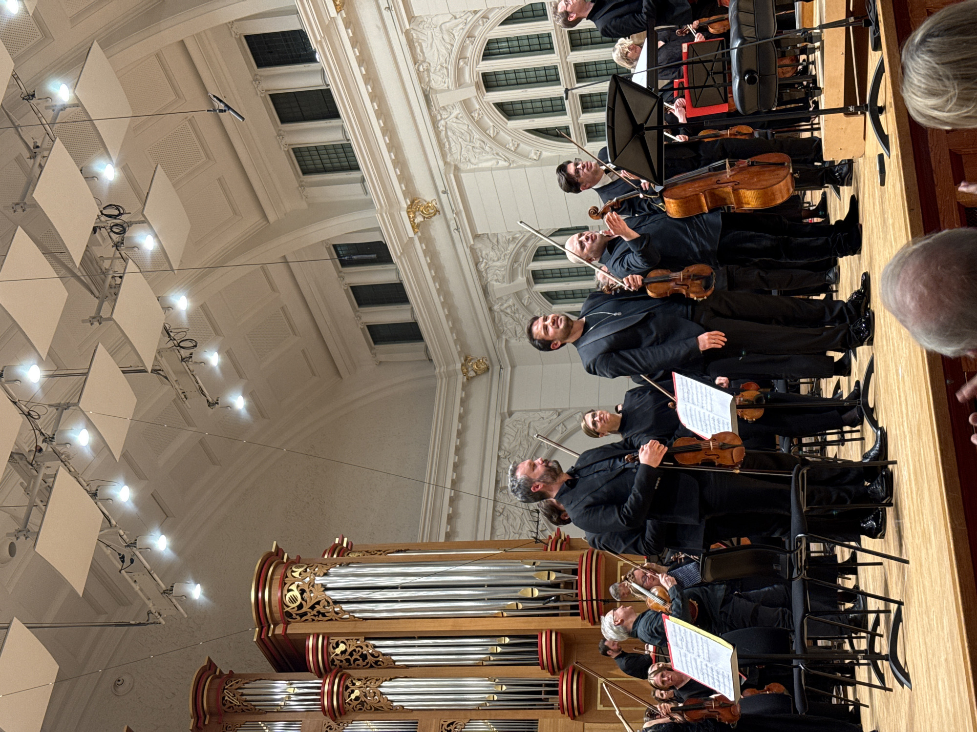 An orchestra performs on stage, likely in London, United Kingdom. An ornate pipe organ stands prominently on the left side of the image. Musicians dressed in black formal attire are seated, holding violins and cellos, with music stands in front of them. The concert hall features an elaborate ceiling with decorative moldings and square light fixtures. The perspective of the photo is from the audience, showing the ensemble from a slightly elevated angle.