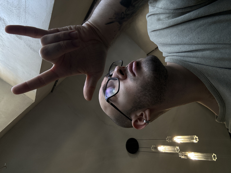 A male individual is depicted indoors, looking upwards and to the right, with his right hand raised in a "corna" (rock on) gesture. He has a shaved head, dark-framed eyeglasses, a beard, and a silver double ear cuff on his left ear. A tattoo is visible on his right forearm. He is wearing a light-colored crewneck t-shirt. The background consists of a light-colored ceiling with a skylight or window providing overhead illumination from the right. To the left, a modern ceiling light fixture with a black circular base and four illuminated, clear, ribbed cylindrical glass pendants hangs downwards. The specific location within Moscow, Russia, is not identifiable from the provided visual information.