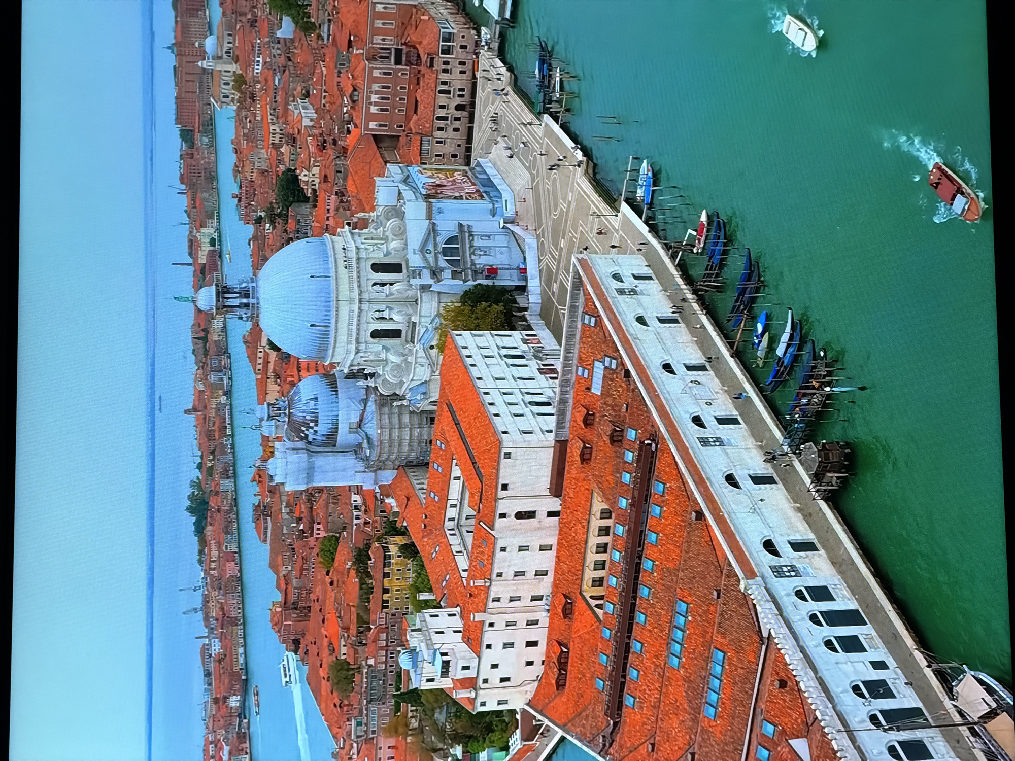 The image shows an aerial view of Venice, Italy, not Chirchik, Uzbekistan. The scene features a prominent white domed church, Santa Maria della Salute, with its light blue dome dominating the center of the frame. The church is surrounded by a dense urban landscape characterized by buildings with red tile roofs. The water is a vibrant turquoise and shows several boats, including gondolas docked along the canals and motorboats navigating the waterways. In the foreground, a long, white building with arched windows lines the waterfront. The sky is a pale blue.