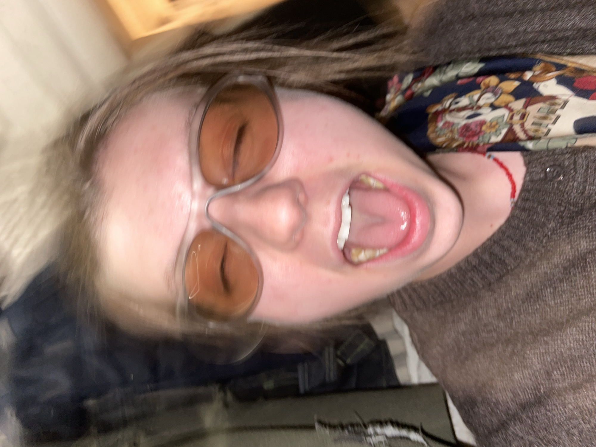 A blurred close-up shot of a person wearing sunglasses with brown lenses, with their mouth open and tongue visible. The person appears to be wearing a brown sweater and a patterned scarf around their neck. A red bracelet is visible on their wrist. The background is indiscernible, making it impossible to determine if the location is Moscow, Russia.