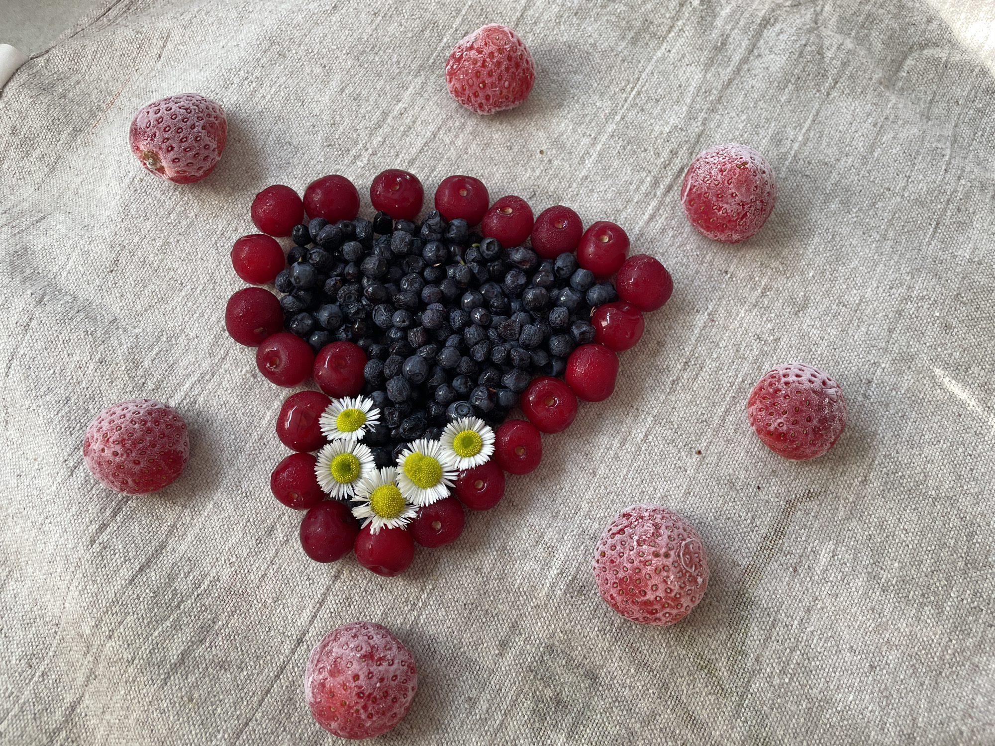 The image displays a heart shape created with blueberries and cherries on a light-colored, textured fabric, likely linen. The heart is outlined by a border of cherries, with a dense cluster of blueberries filling the center. Several small white and yellow flowers are placed within the blueberry cluster. Scattered around the heart shape are several frozen strawberries, each covered in a layer of frost. The overall composition suggests a deliberate arrangement of food items, possibly for decorative or culinary purposes. The location in Brest, Belarus, is not discernible from the image itself.