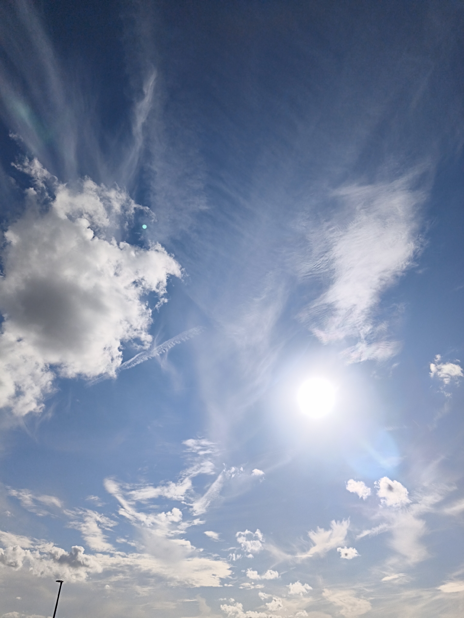 The image depicts a bright, sunny sky with scattered clouds over Tallinn, Estonia. The sun is a prominent feature, shining intensely and creating lens flare effects. Wispy, cirrus clouds are spread across the deep blue sky, interspersed with larger, cumulus clouds. In the lower part of the image, a dark, thin pole or mast is visible, extending upwards from the bottom edge. The overall scene is one of a clear day with dynamic cloud formations.