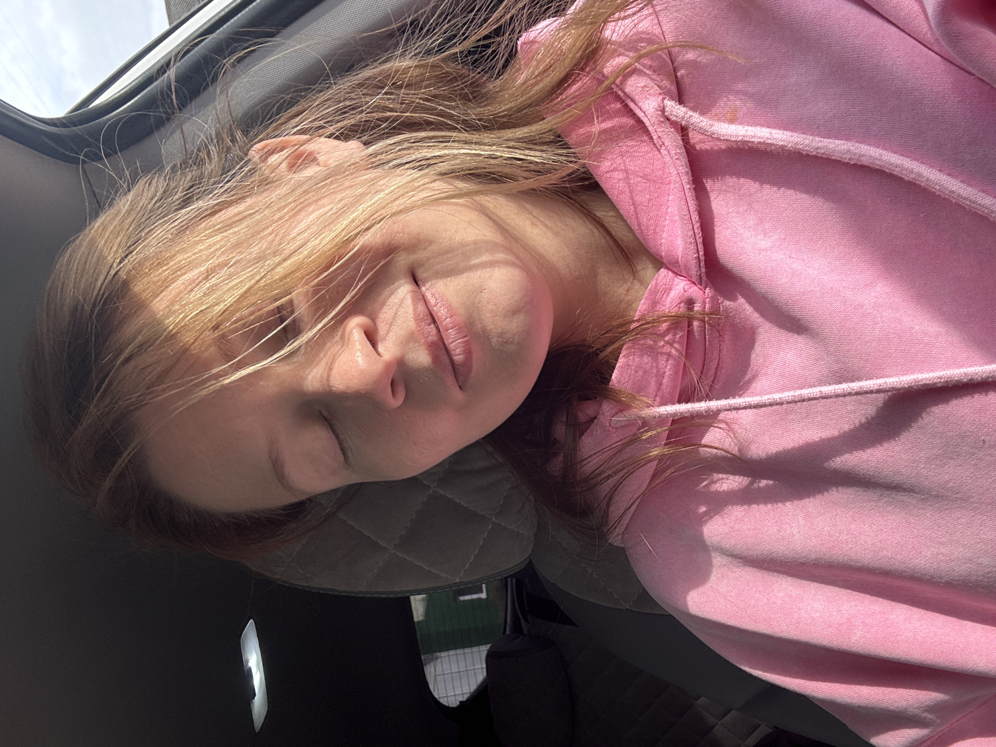 The image, rotated 90 degrees clockwise, captures a young woman resting inside a vehicle, likely a car, in Chaikovsky, Russia. She has long, light brown or blonde hair and is wearing a bright pink hooded sweatshirt. Her head is tilted and resting on a gray, quilted car headrest.
The woman appears to be sleeping or deeply relaxed, with her eyes closed and a gentle, peaceful smile on her lips. Direct, strong sunlight illuminates her face and clothing, casting distinct shadows across her features and the folds of her hoodie, suggesting it is daytime and sunny. A sunroof is partially visible at the top of the frame, confirming the indoor automotive setting.
Notable details include the texture of the pink hoodie, the drawstring, and the quilted pattern of the headrest. A small, lighter-colored mark is visible on the pink fabric near the hood's seam. The overall impression is one of tranquility and comfort during a journey or a break. No discernible text is visible in the image.